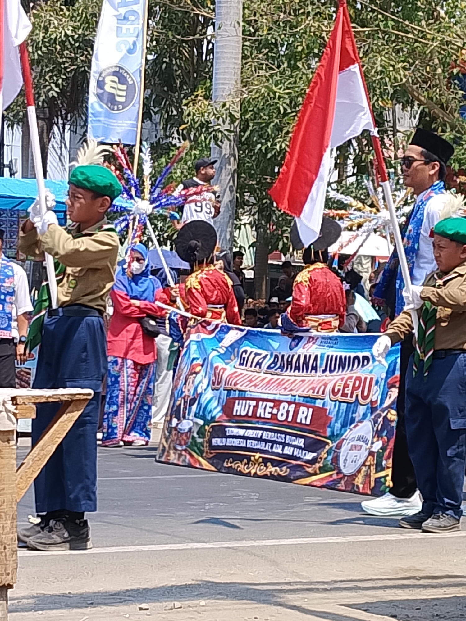 The image depicts a street scene in Balun, Indonesia, likely during a parade or cultural event. In the foreground, two young boys in Scout-like uniforms, complete with green berets and neckerchiefs, hold flags. One boy on the left holds a white flag with a red stripe at the top. The boy on the right is partially obscured by a large banner. The banner features text in Indonesian, including "GITA BAHANA JUNIOR MUHAMMADIYAH CEPU" and "HUT KE-81 RI," suggesting a celebration of the 81st anniversary of Indonesian Independence by a junior musical group from Muhammadiyah Cepu.
Behind the Scouts, several individuals are visible. A woman wearing a blue hijab and a red top stands with her arms crossed. To her right, two people in ornate red and gold costumes appear to be part of a performance. Further in the background, a crowd watches the proceedings, some holding umbrellas. A man wearing a black peci and sunglasses stands near the Indonesian flag.
The environment suggests a sunny day with trees lining the street. The ground is asphalt with white markings. The overall atmosphere is festive and community-oriented.