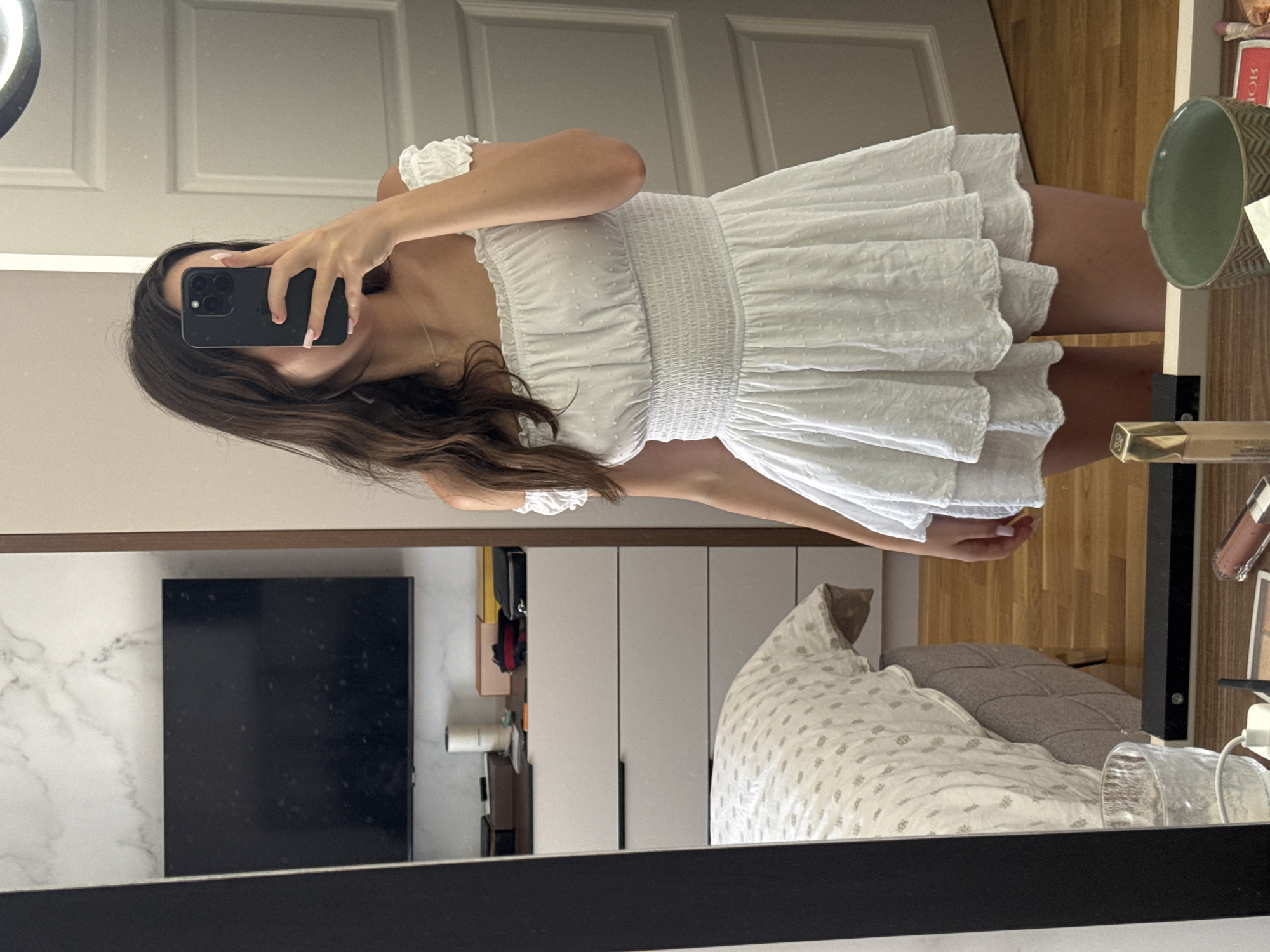 In an indoor setting, a young woman with long, dark hair stands in front of a mirror, taking a photo of herself with a smartphone. She is wearing an off-the-shoulder white mini-dress with smocking around the bodice and a tiered skirt. The dress has a textured pattern of small dots. The background shows a bedroom with a bed covered in a white patterned comforter, a dresser with several drawers, and a television mounted on a marble-patterned wall. A large mirror reflects the scene, and on a surface in front of the mirror, several makeup items are visible, including a lipstick and a bottle of foundation. The lighting appears to be natural, coming from a window or overhead light source.