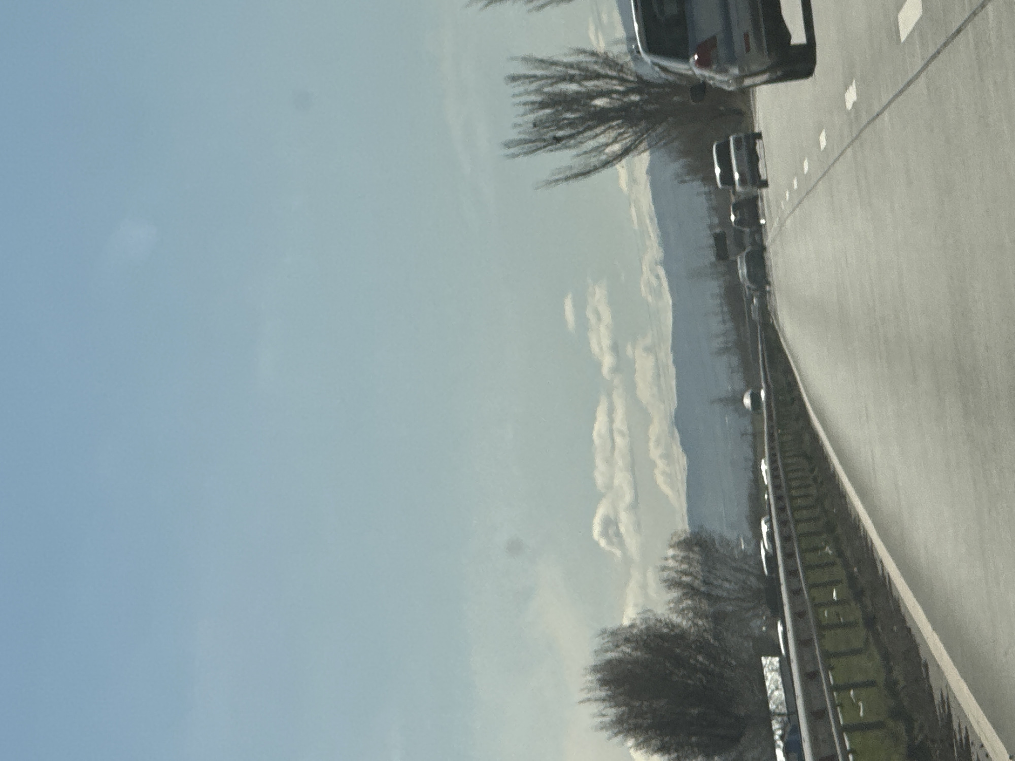The image shows a view from a vehicle traveling on a highway in an unknown location. The sky is blue with some clouds visible. A mountain range is present in the distance. The highway has multiple lanes, and vehicles are seen traveling in the same direction. A guardrail separates the highway from a grassy area. Trees are present on the side of the road.