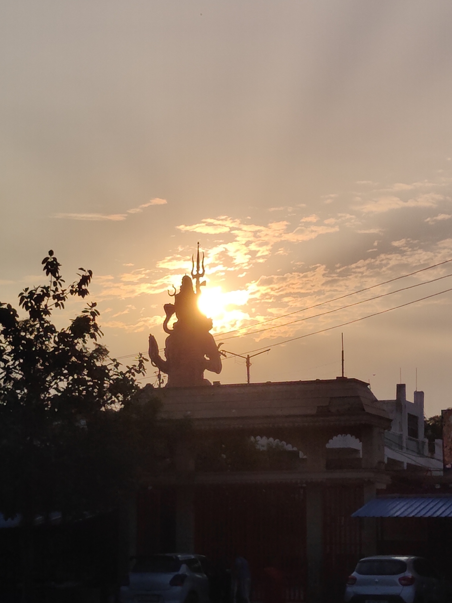 The image depicts a silhouetted statue of Lord Shiva, complete with a trident, against a sunset sky in Gurgaon, India. The sun is partially obscured by the statue, casting a bright glow. In the foreground, a leafy tree is visible on the left, and a building structure with an arched entrance and a pitched roof occupies the middle ground. Utility wires are strung across the sky. In the lower portion of the image, two cars are parked, one silver hatchback on the right and another partially visible car on the left. The overall scene captures a serene evening atmosphere with a prominent religious monument.
