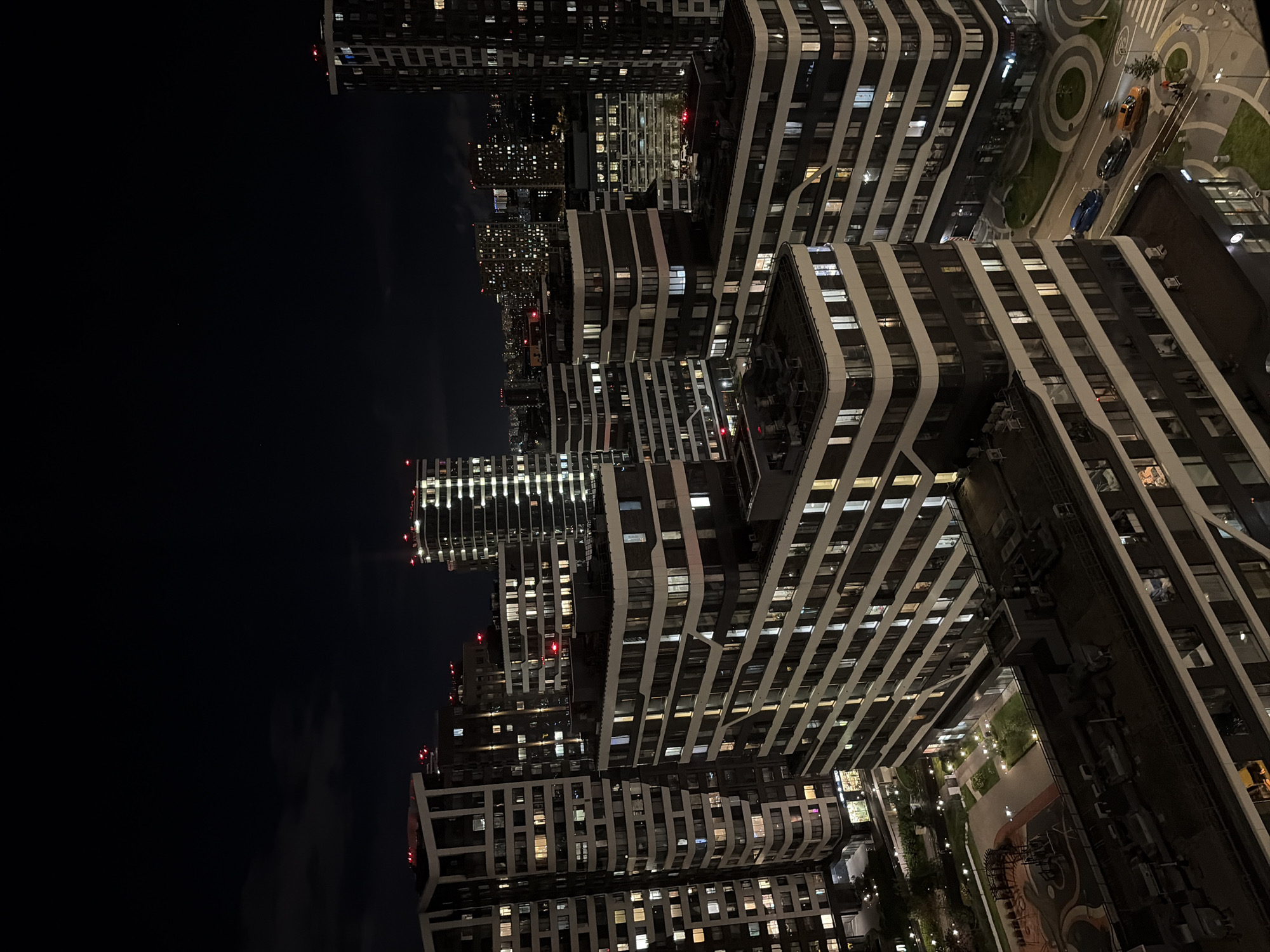 The image depicts an aerial view of a residential complex at night in Moscow, Russia. Multiple modern, multi-story buildings with numerous illuminated windows dominate the scene. The architecture features contemporary designs with curved facades and dark exterior paneling contrasted with white or lighter-colored horizontal banding. Several buildings are tall and slender, while others are wider and more block-like.
In the foreground, the roofs of some buildings are visible, some with visible HVAC units. Below, a courtyard area with a playground, grassy patches, and paved walkways is illuminated by streetlights. A road runs alongside the complex, with several cars, including a blue sedan, a black car, and an orange car, visible on it. Pedestrians can be seen on a crosswalk.
The sky above is dark, with some faint clouds and distant city lights suggesting a sprawling urban environment. The overall impression is one of a well-developed, modern residential area functioning at night. The lighting emphasizes the architecture and the lit interiors of the apartments, creating a sense of activity within the buildings. The image captures the characteristic nighttime ambiance of a modern city district.