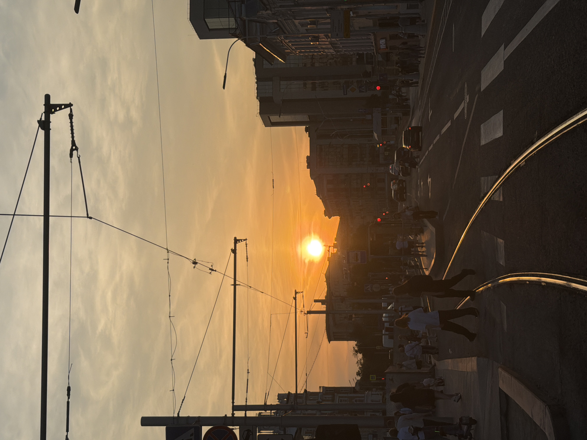 This is an urban street scene captured during sunset in Moscow, Russia. The specific location is **Myasnitskaya Street**, with the camera facing west.
The sky is dominated by a vibrant orange and yellow hue, intensifying around the low-hanging sun, which appears as a brilliant, almost white disc, positioned slightly left of the image center. The strong backlighting creates deep silhouettes of most subjects.
In the immediate foreground, a set of double tram tracks curves from the lower-left to the mid-right. The rails reflect the golden sunlight, appearing as bright, thin lines on the otherwise dark asphalt road. A white pedestrian crossing (zebra stripes) is visible on the road surface to the right of the tracks. The road itself appears multi-laned.
On the left side of the foreground, a dark-silhouetted figure is seen on an electric scooter on a sidewalk. Numerous pedestrians are present on both sidewalks and crossing the street in the mid-ground. Two prominent figures, one in a lighter-colored top and the other in dark attire, are captured mid-stride crossing the tram tracks in the central foreground.
Overhead, a dense network of black electrical and tram wires, supported by various poles and gantries, crisscrosses the upper half of the frame, sharply silhouetted against the bright sky. A street sign with a red circular border and a diagonal line (typically indicating "no stopping") is partially visible on a pole on the far left.
Along the street, buildings line both sides. On the left, multi-story buildings with traditional, possibly ornate, architectural features are visible in silhouette. Towards the center-right and in the background, several taller, more modern buildings with reflective glass facades stand out, also largely silhouetted. On the far right, an older, multi-story building with distinct window patterns and cornices is visible; a partially legible sign with Cyrillic characters, possibly "РЕДПОСТ" (Redpost), is visible on its facade in the lower portion of the building.
Multiple red traffic lights are visible throughout the mid-ground, indicating current stop signals or turn restrictions. Several cars are present on the road in the mid-ground, appearing as dark silhouettes with some visible red taillights. The overall atmosphere conveys the end of a day in a bustling city.