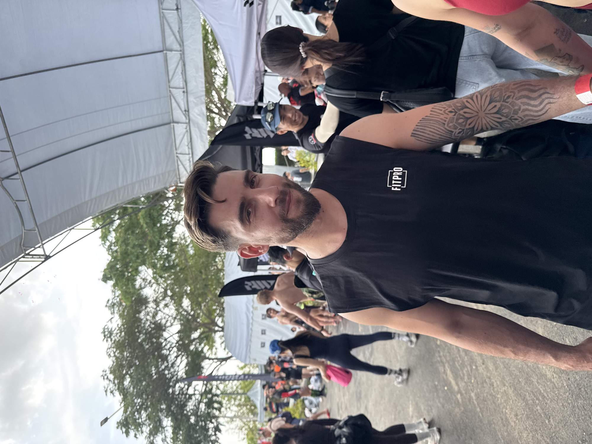 A medium shot captures a man with dark hair and a beard, wearing a black t-shirt with "FITPRO" printed on it. He is looking towards the camera with a slight smile. A portion of a tattoo is visible on his right arm. Behind him, there are tents, people, and trees. The setting appears to be a gathering or event with individuals performing exercises, possibly a fitness event in บ้านสำนักตะแบก, Thailand. Some are doing push-ups. A woman with a tattoo on her arm is also in the shot.