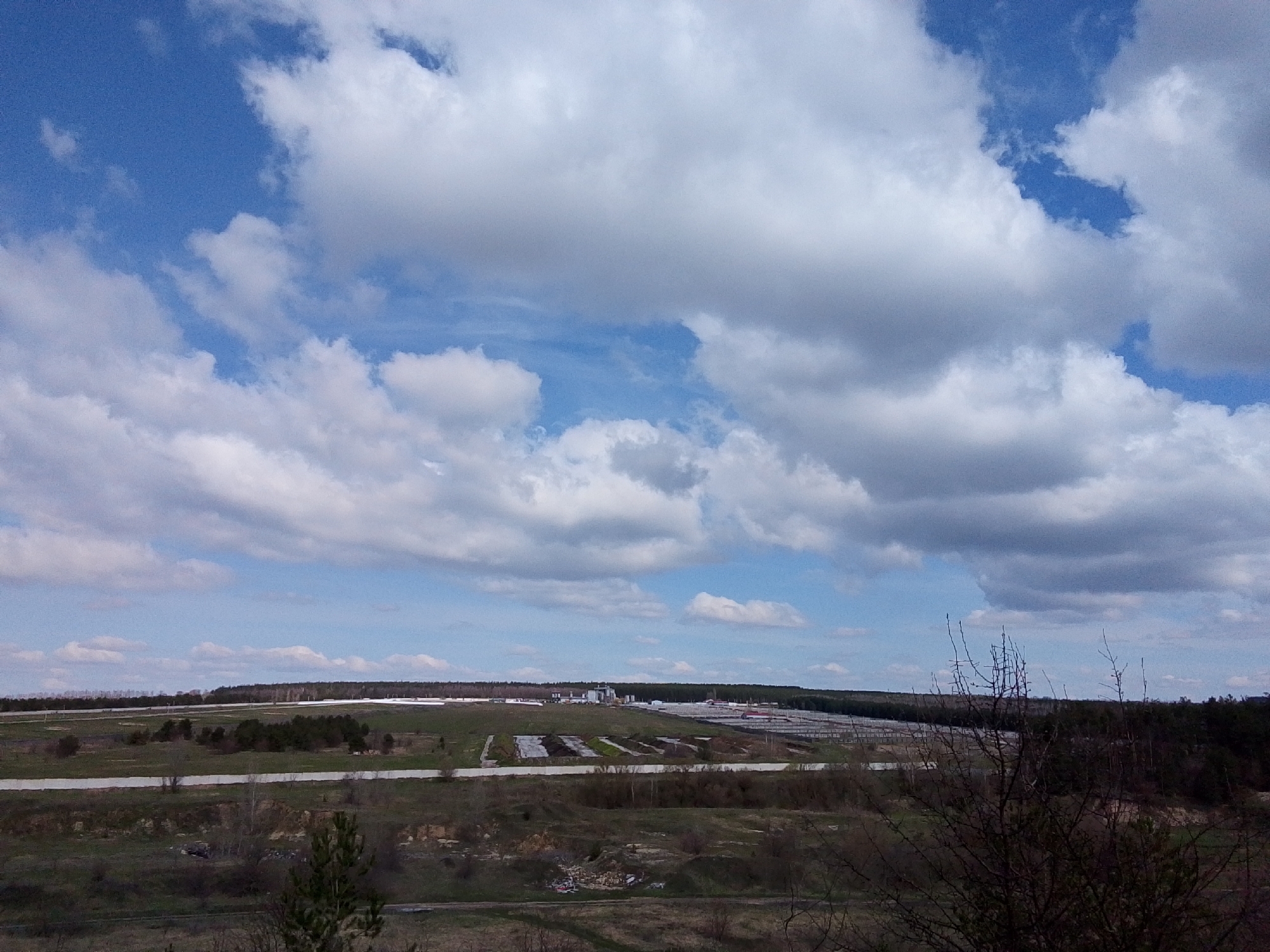 This panoramic landscape photograph, taken in Khlevnoye, Russia, captures a vast outdoor scene under a dynamic sky. The upper half of the image is dominated by a mix of bright blue sky and large, fluffy white and grey clouds, suggesting a partly cloudy day.
The foreground consists of rugged, uneven terrain with sparse dry grass, bushes, and a few leafless tree branches, particularly noticeable on the right side. A dirt path or road is faintly visible cutting through this area.
The middle ground transitions into a flatter expanse of land featuring patches of green vegetation and several long, low-lying structures, likely industrial or agricultural buildings such as greenhouses or animal enclosures, characterized by their white or light-colored roofs. In the central middle distance, a cluster of taller industrial buildings, possibly silos or processing facilities, stands out against the horizon. Several light-colored horizontal strips, which could be unpaved roads or tracks, traverse this area, connecting different sections of the site.
The background is defined by a dense line of dark green forest stretching across the horizon, separating the cultivated or industrial area from the sky. The overall lighting suggests it is daytime, possibly late morning or early afternoon, with natural light illuminating the landscape. There are no discernible human figures or specific activities taking place, and no visible text. The scene presents a blend of natural landscape and human-made agricultural or industrial infrastructure.