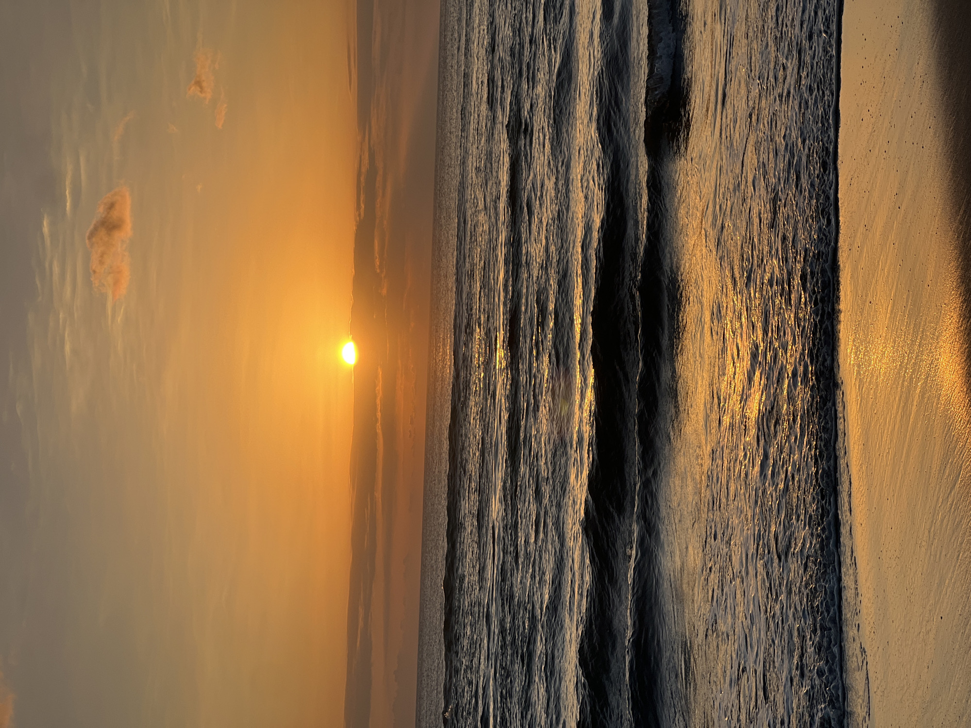 This image captures a serene sunset over the ocean, likely at Kuta Beach in Bali, Indonesia. The sun, a bright orange orb, is partially obscured by a thin layer of clouds near the horizon, casting a warm golden glow across the sky. The sky itself is a gradient of orange and lighter yellow, with scattered wispy clouds tinged with pink.
The ocean in the foreground is a deep, dark blue, with gentle waves rolling towards the shore. The sunlight reflects off the water's surface, creating shimmering streaks of gold. The waves are capped with white foam as they reach the wet sand. The beach in the very bottom of the frame is a light brown, wet sand, reflecting the sunset's light.
There are no people or specific objects in the scene, focusing solely on the natural beauty of the sunset and the seascape. The overall impression is one of tranquility and natural splendor.