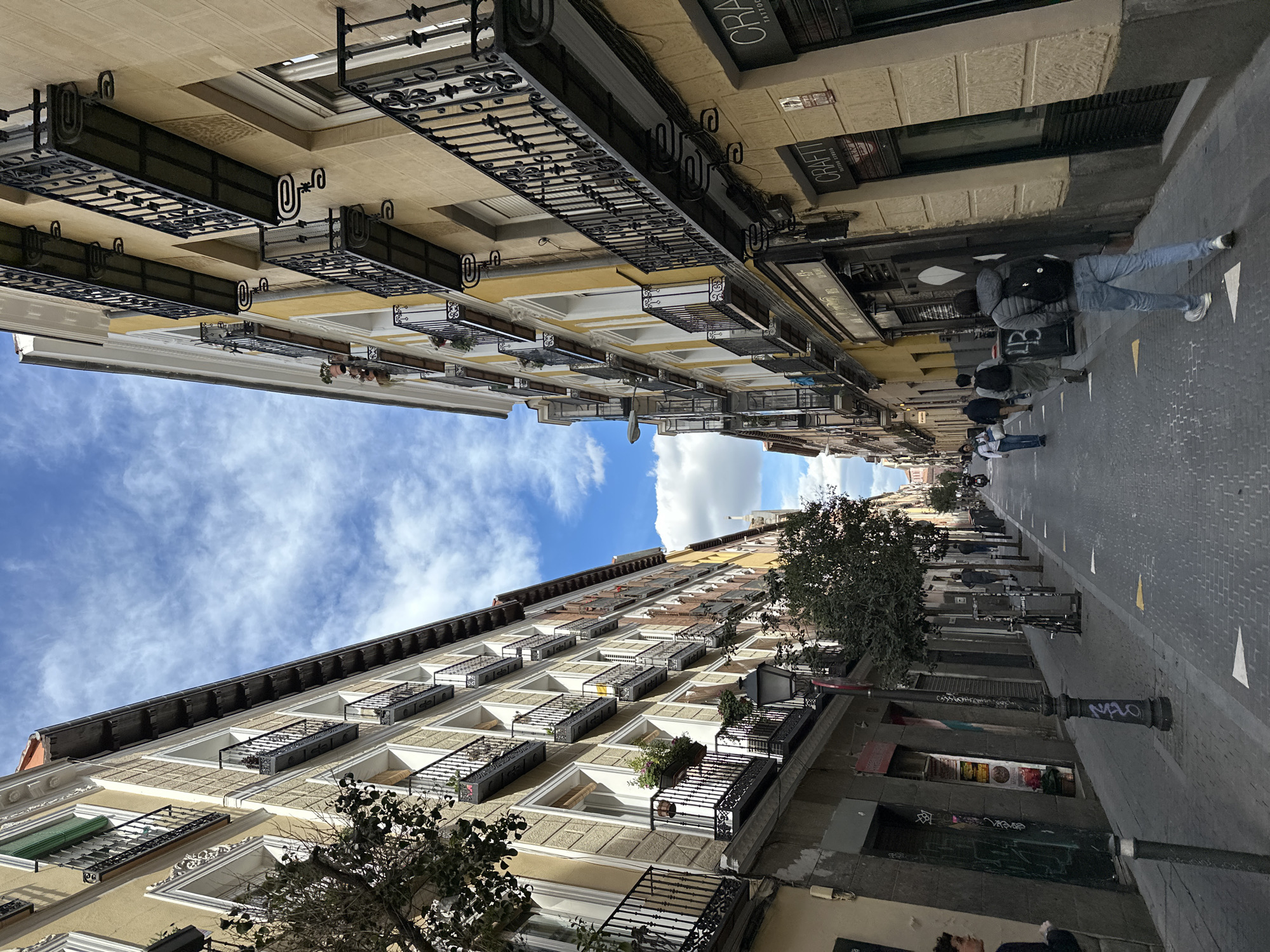 The image depicts a street in Madrid, Spain, viewed from a low angle, almost directly upward. The street is narrow, flanked by multi-story buildings with wrought-iron balconies. The buildings are characterized by classical architectural details and beige/yellow facades. Pedestrians are visible walking along the right side of the street. There are trees lining parts of the street, and the sky is visible between the buildings, showing a blue sky with scattered clouds. Graffiti is visible on some of the lower building facades. The street appears to be paved with cobblestones or similar materials.