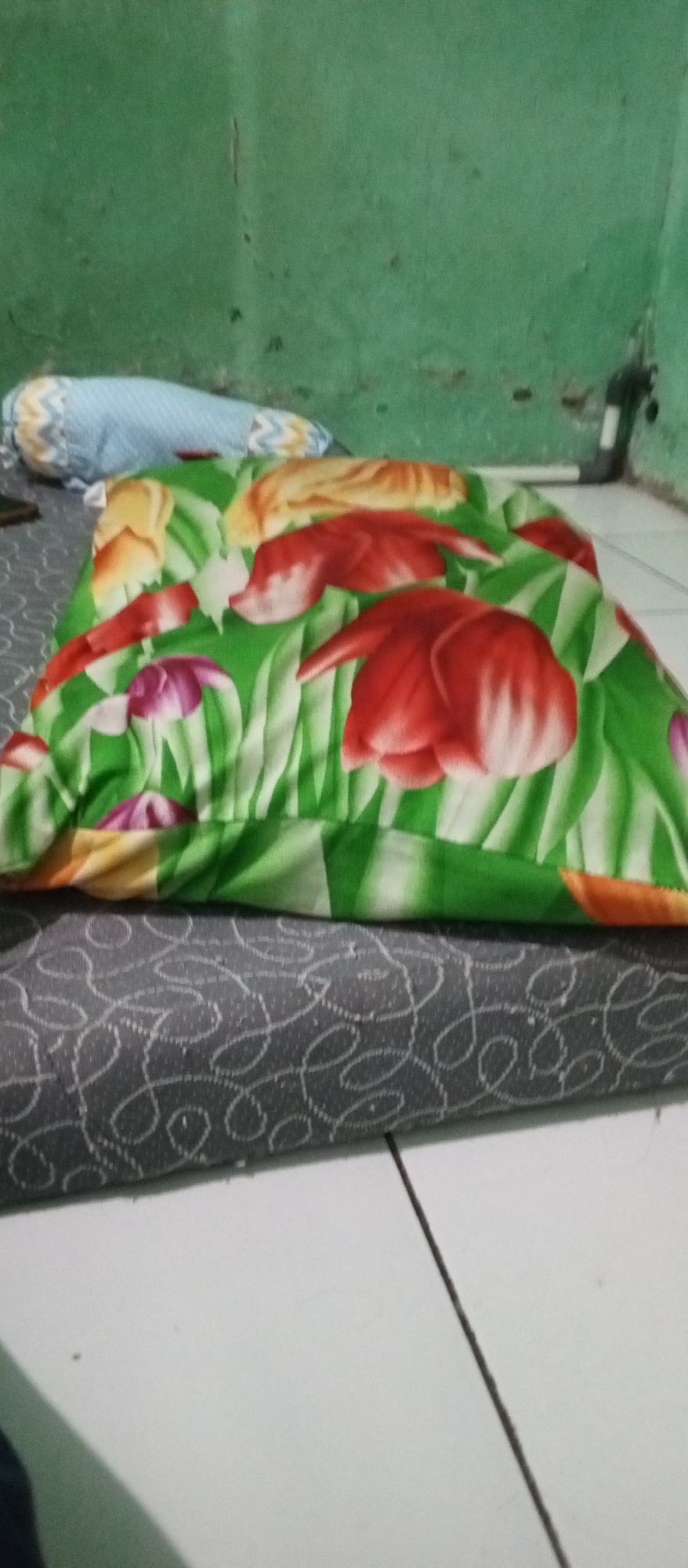 The image shows a bed with a floral patterned pillow and a striped bolster pillow, placed against a green wall. The bedding appears to be on a mattress which is on the floor. The floor is tiled and a dark object is visible in the bottom left corner of the image. The scene is likely indoors, possibly in a residential setting in Karawang, Indonesia.