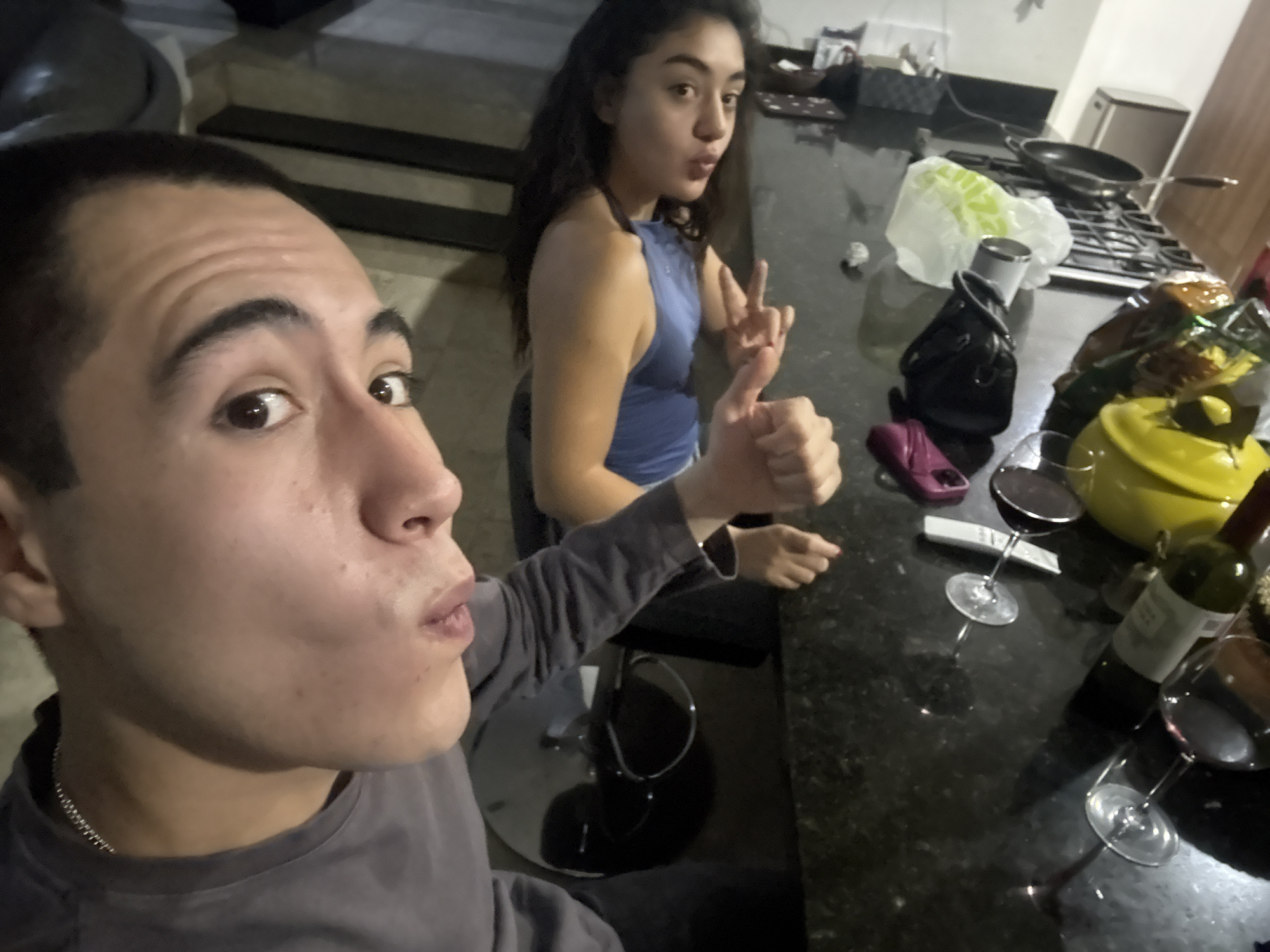 A close-up selfie shows a young man with short dark hair wearing a grey shirt and silver chain, holding his thumb up. Beside him, a young woman with long dark hair, wearing a blue halter top, is making a peace sign. They are seated at a dark countertop, which holds various items: two filled wine glasses, a bottle of red wine, a yellow teapot, a black purse, a cell phone, and other unidentified objects. In the background, a stove with a pan, a garbage bag, and kitchen cabinets are visible. The location is likely a private residence, possibly in Santa Clara Ocoyucan, Mexico.