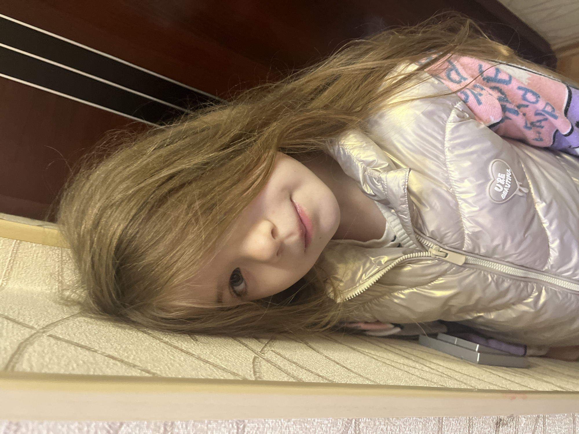 A young girl with blonde hair is lying on a surface with a quilted pattern. She is wearing a shiny, light-colored jacket with a pink and purple patterned garment visible beneath it. The jacket has a logo that reads "U'RE BEAUTIFUL." A dark brown cabinet with light-colored stripes is visible in the background. A desk or table is visible in the bottom of the image. The location is not identifiable from this image.