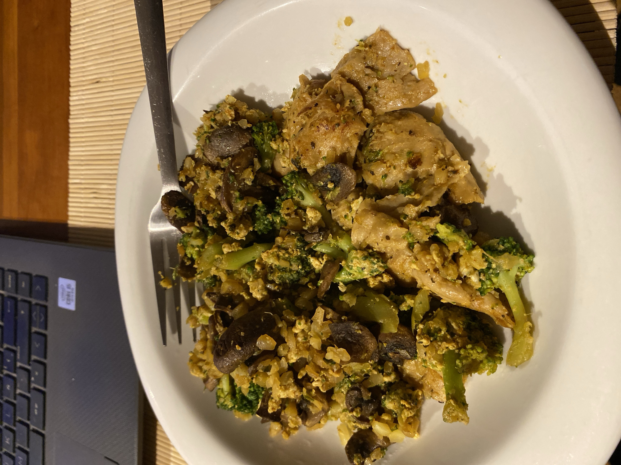 The image displays a close-up, top-down view of a meal served on a white, round plate, suggesting an indoor setting. The plate is filled with a robust dish primarily composed of cooked chicken pieces, identifiable by their light golden-brown color and textured surface. Mixed with the chicken are bright green broccoli florets and dark, sliced mushrooms. The base of the dish features a yellowish, crumbly texture, possibly indicating rice or cauliflower rice combined with egg or a sauce, creating a rich, flavorful appearance. A silver-colored fork rests on the left side of the plate, with its tines partially submerged in the food, indicating that the meal is either about to be eaten or is already in progress.
To the left of the plate, a dark grey laptop keyboard is visible, featuring several keys and a clear sticker with the "Intel Core i5" logo. A light-colored placemat, possibly woven bamboo, is partially visible beneath the plate and laptop, resting on a warm-toned wooden surface, likely a table. The lighting appears warm and somewhat dim, consistent with an evening meal enjoyed indoors. The overall scene suggests a casual dining experience, possibly while working or relaxing with a laptop nearby. No individuals are visible in the image. While the location context of Gijón/Xixón, Spain, is provided, there are no visual cues within the image itself to confirm this.