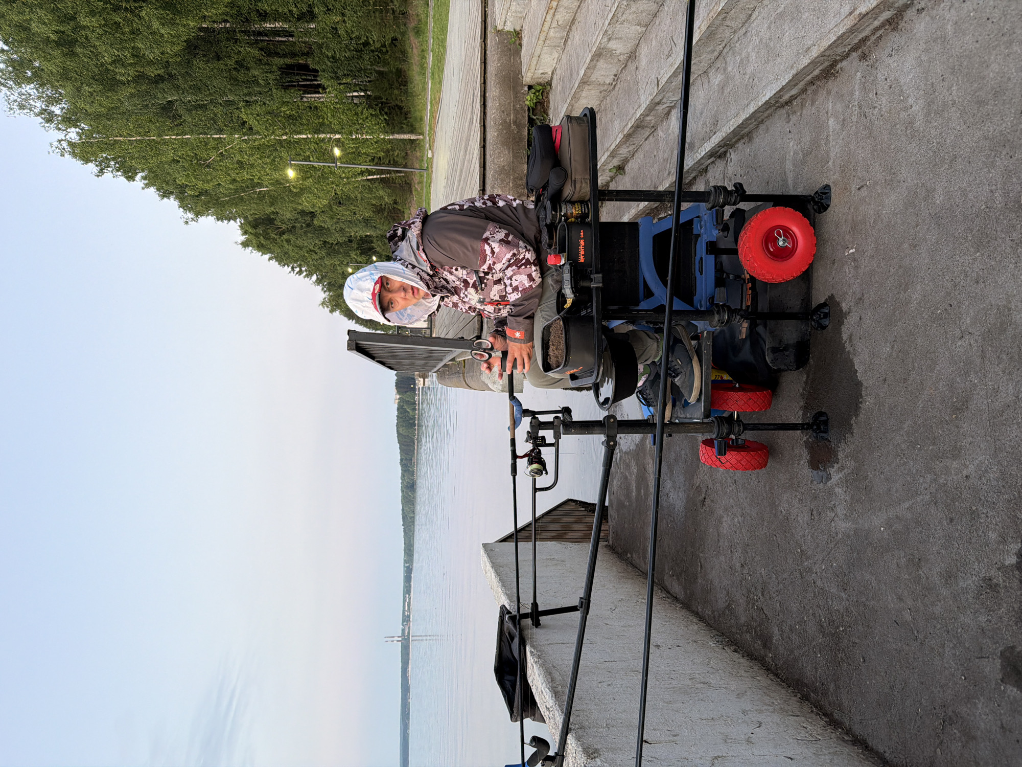 A person sits in a blue fishing chair with red wheels on a concrete pier by a body of water. The person wears a camouflage jacket, a white hooded hat, and dark pants. The person holds a pair of scissors in their right hand. In front of the person is a black tray with various fishing items, including containers of bait and small jars. The background shows a calm body of water, a distant industrial structure with two smokestacks, a line of green trees, and some outdoor lighting fixtures. The scene appears to be a waterfront area, possibly in Chaikovsky, Russia, given the presence of industrial elements often associated with the city.