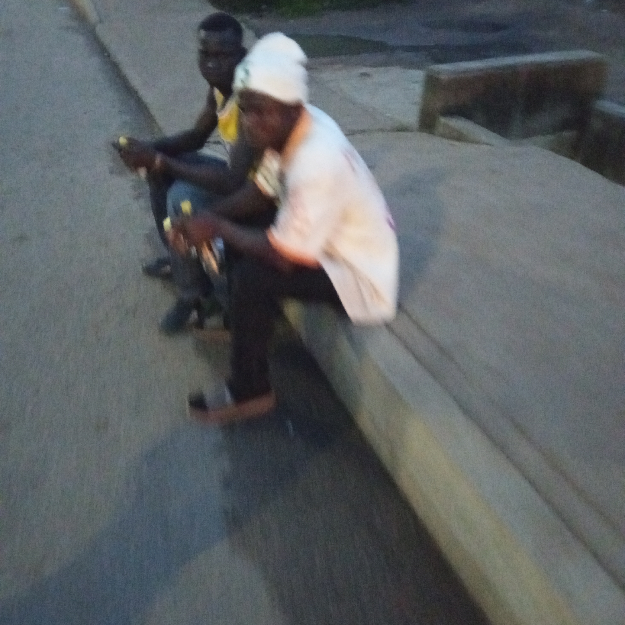 Two individuals are seated on a raised concrete curb at the side of a paved road. The person on the right is wearing a white head covering and a white long-sleeved shirt, and appears to be holding something yellow in their hands. The person on the left is wearing a yellow shirt and dark trousers. The scene is outdoors in what appears to be a city environment, potentially Bukuru, Nigeria, indicated by the context provided. The lighting suggests it might be dusk or early evening. The background shows a blurry concrete structure and a road surface. The image is somewhat out of focus, particularly the faces of the individuals.