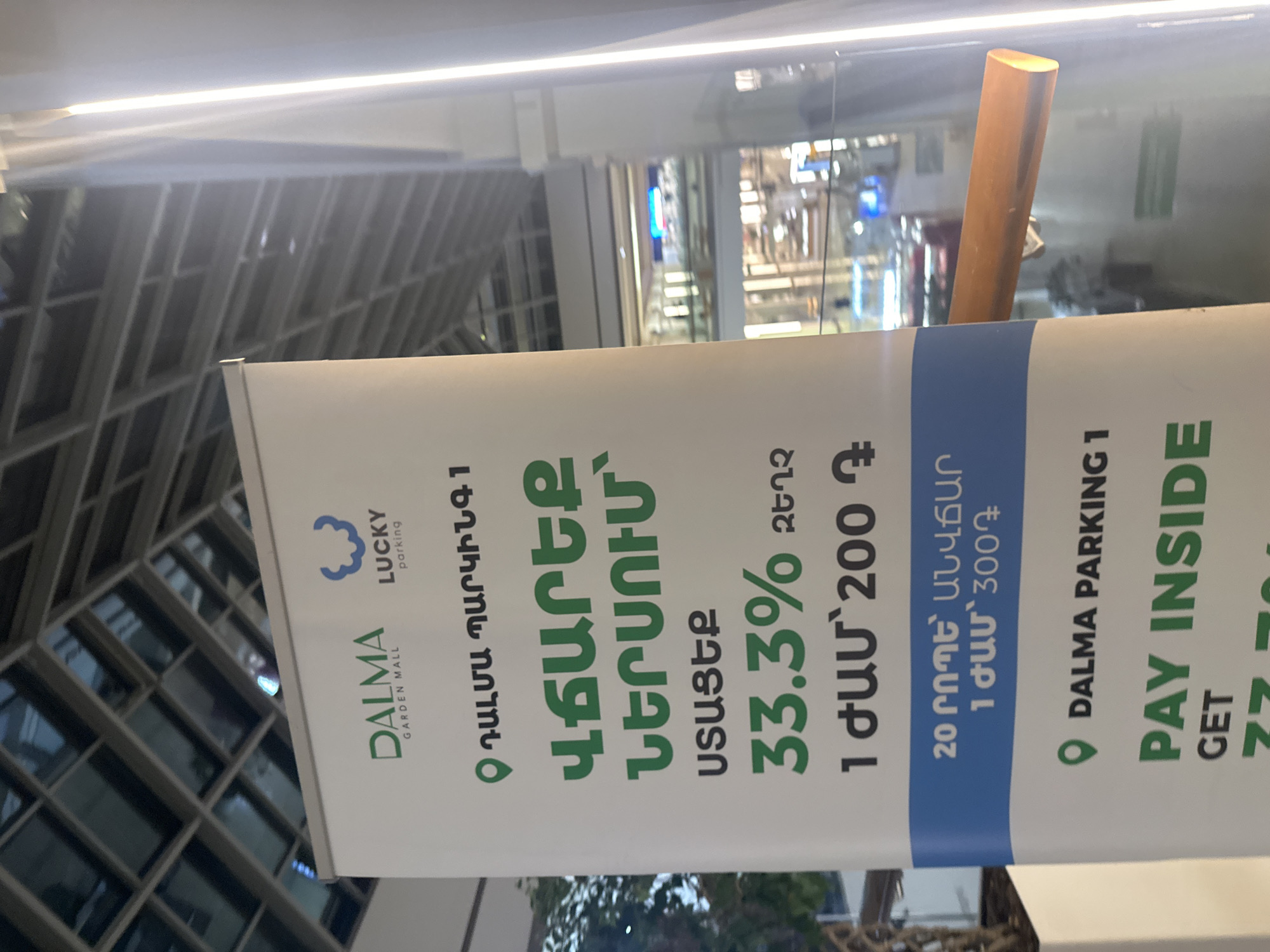 The image shows a sign advertising parking at Dalma Garden Mall in Yerevan, Armenia. The sign is white and green, with a blue section. It lists prices and discounts in Armenian and English.