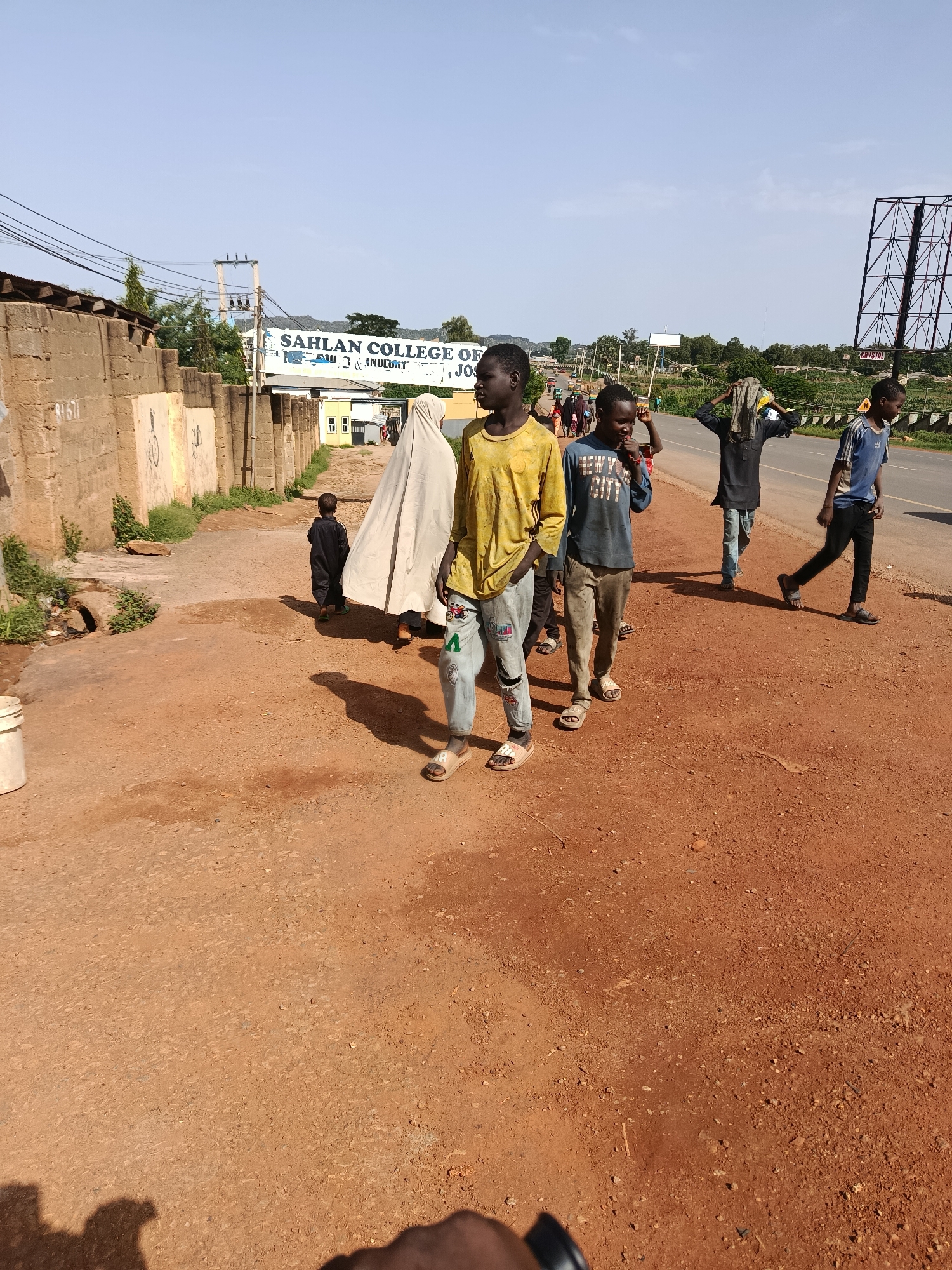 A group of individuals walk along a dirt road next to a highway under a clear sky. A billboard is visible in the distance. In the foreground, a person's hand and arm are partially visible, as if holding the camera. A child in black clothing walks in the middle ground, with a person in white covering their head and body behind them. Several boys walk towards the highway, one in a yellow shirt, another in a blue shirt with text, and another in a blue t-shirt. A person in a black shirt with a cloth on their head walks behind them. The environment appears to be a roadside in a warm climate, with red dirt covering the ground. A brick wall runs along the left side of the road.