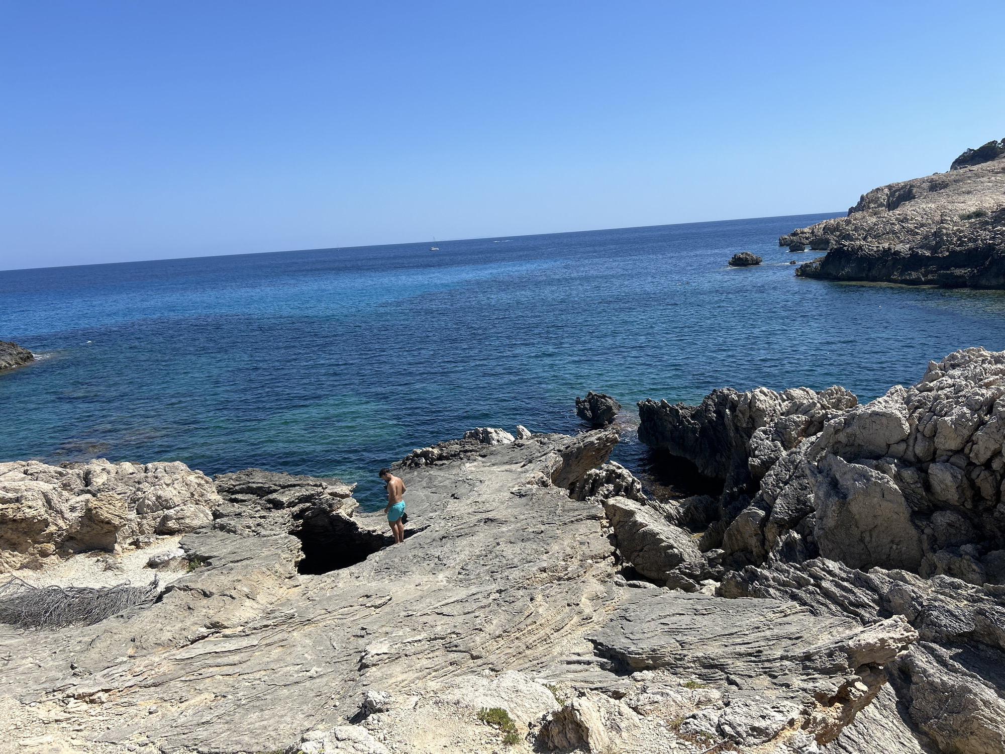 A person in blue swim trunks stands on a rocky shoreline with the ocean in the background. The sky is clear blue and the water is a deep blue with some turquoise hues. The rocks are rough and uneven, with some vegetation growing in between them. The person is looking down at the rocks, and appears to be walking. There are some smaller rocks and cliffs in the distance, as well as some faint boat shapes on the horizon. The overall scene is serene and natural.