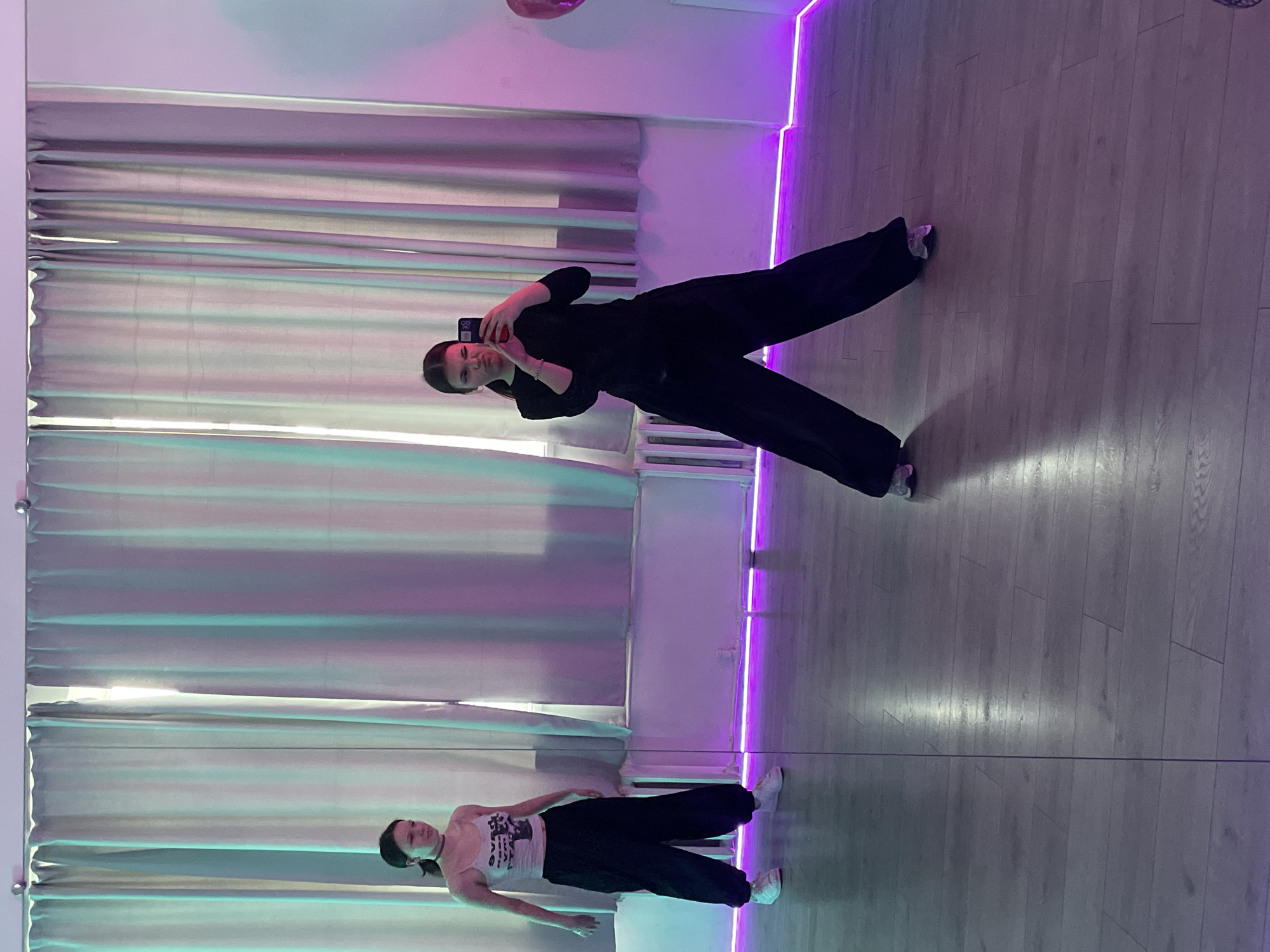 A person wearing a black outfit is taking a selfie in front of a large mirror in a room. The reflection in the mirror shows another person wearing a light-colored shirt and dark pants. Both individuals are inside a room with curtains on one side, a light strip running along the wall, and a wooden floor. While the exact location within Saint Petersburg, Russia is unknown, the scene suggests a dance studio or similar space based on the presence of the mirror and dance-like attire.
