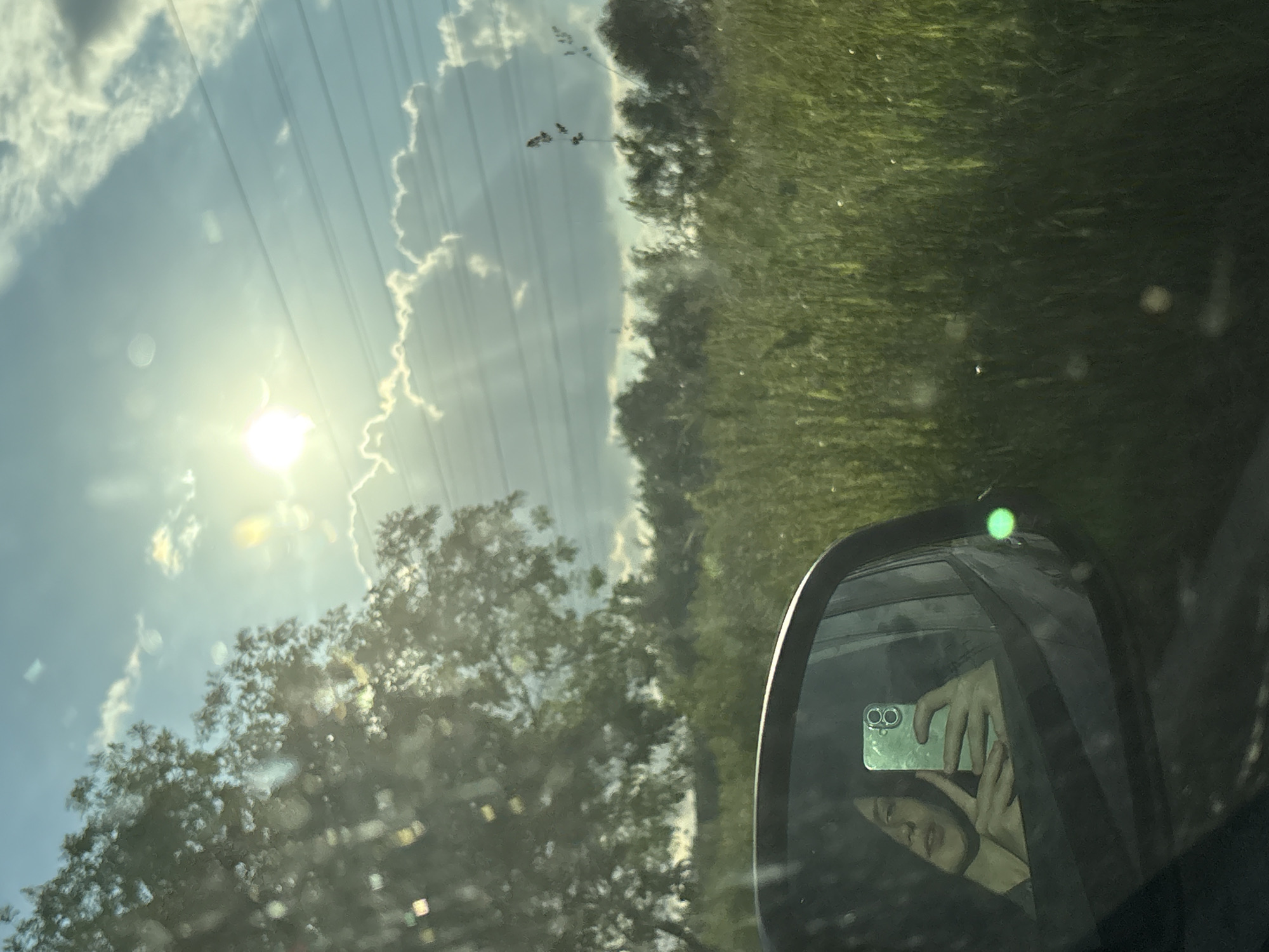 A selfie is taken in the side mirror of a vehicle, reflecting a young person with dark hair holding a smartphone. The person is making a peace sign with their fingers. The background visible in the mirror and outside the vehicle shows a lush, green field with tall grass and trees under a bright, partly cloudy sky. Sunlight streams through the clouds, creating lens flare and light streaks. The setting appears to be rural or on the outskirts of a city. There is no indication that this location is in Saint Petersburg, Russia.
