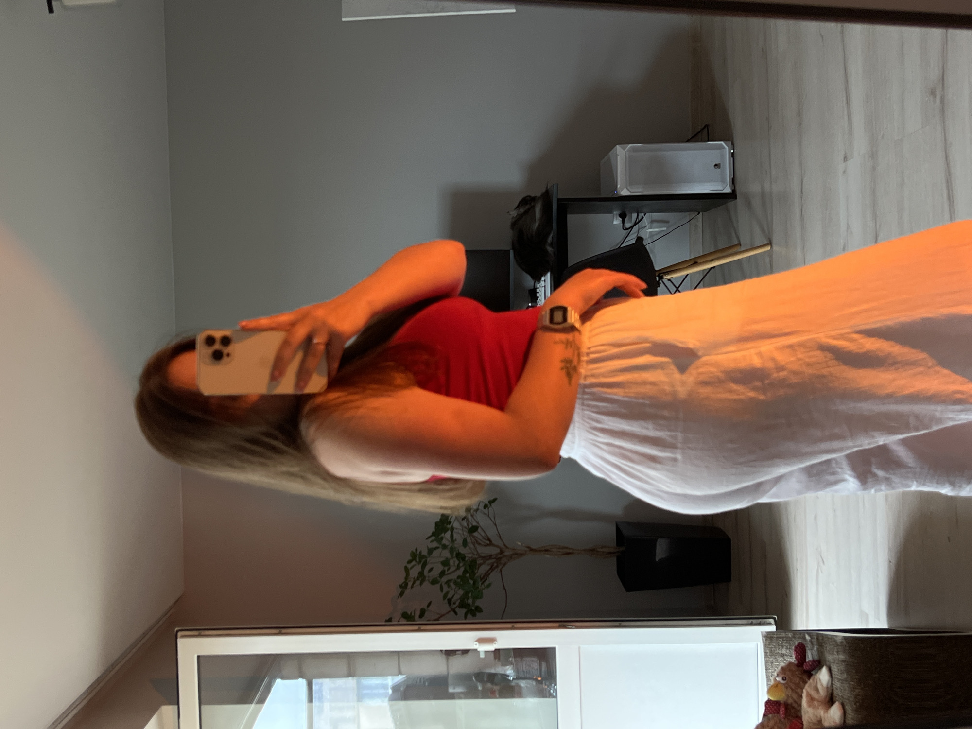 The image, taken in Ternopil, Ukraine, shows a person taking a mirror selfie indoors. The subject is a person, likely female, with long brown hair, wearing a red sleeveless top and a flowing white skirt or wide-leg trousers. She holds a light-colored smartphone up to her face, obscuring most of her features, and wears a digital watch on her left wrist and a ring on her left hand.
The setting appears to be a contemporary room, possibly a living room or bedroom. The walls are painted a light grey, contrasting with a light-toned wooden floor. In the background, to the right of the person, a black shelving unit is visible, holding a white computer case or similar electronic device, with some cables hanging down. Beneath the shelves, a small green plant in a dark pot sits on the floor.
Natural light seems to illuminate the scene, casting warm tones, particularly on the person's white skirt and skin. In the bottom right corner, a glimpse of decorative items, including what appears to be a plush rooster or chicken, can be seen resting on the floor or a low surface. The overall impression is a casual, everyday moment captured in a well-lit indoor environment. No visible text is discernible in the image.