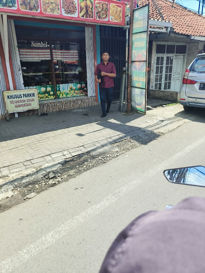 The image was taken from the perspective of a person on a motorcycle, facing forward. The motorcycle is parked on the edge of a paved road with a white line down the middle. The road is in poor condition, with several cracks and patches. To the left is a sidewalk made of concrete pavers. To the right is a building with a tiled roof, a green door, and a white car parked in front of it. The car is facing away from the viewer and is partially obscured by the building. A man is walking on the sidewalk towards the left, carrying a dark bag in his right hand. The man is wearing a red button-down shirt, dark jeans, and brown shoes. The building in the background has an awning with the word "Sambel" on it, and a sign with the words "KHUSUS PARKIR WARUNG NASI BAKSO" on it. The coordinates indicate a location in Padalarang, Indonesia.