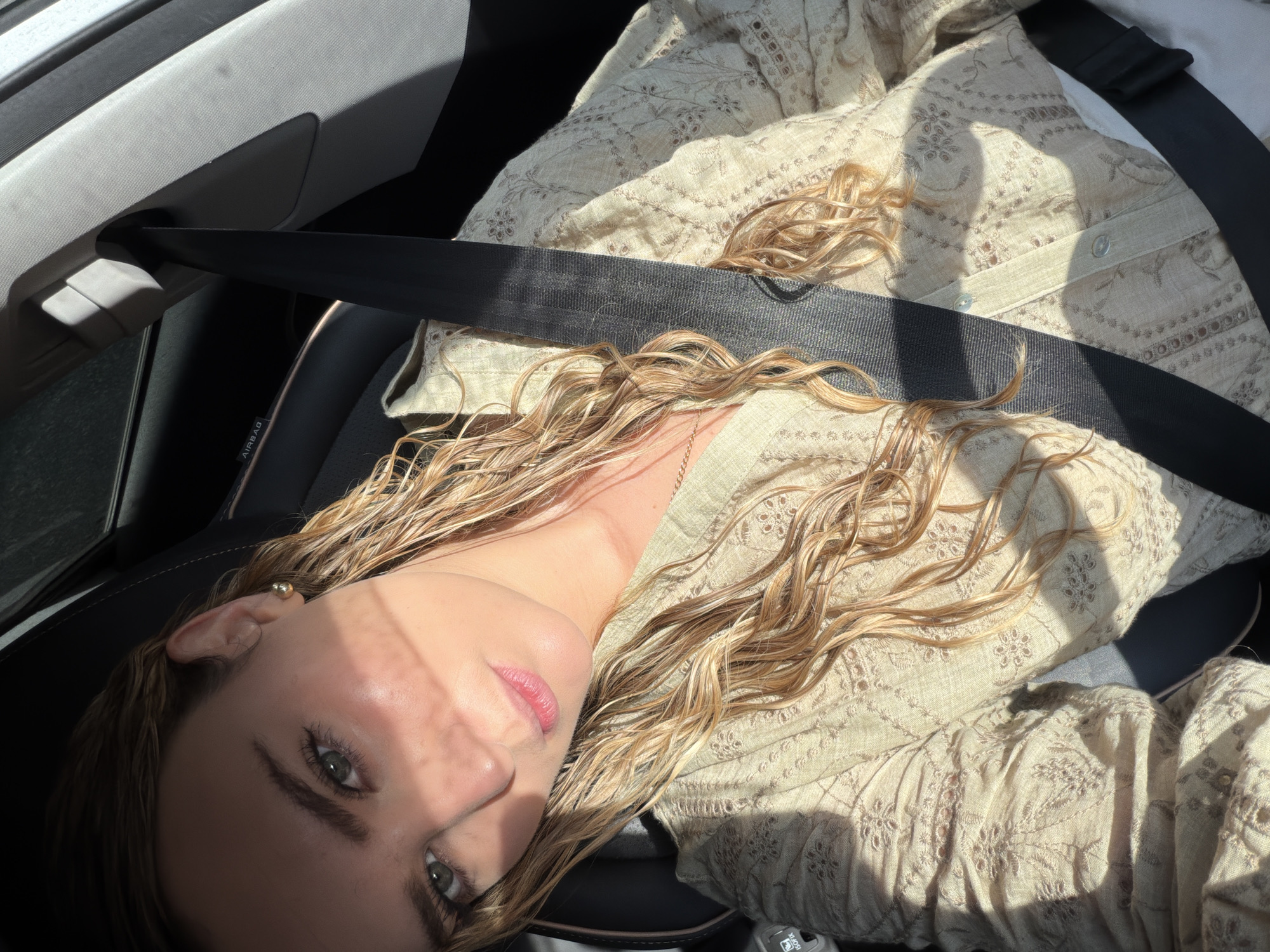 A woman with blonde, wavy hair is in a car. She is wearing a beige patterned shirt and has a black seatbelt across her chest. The environment appears to be the interior of a vehicle. There is an "AIRBAG" label visible. The city of Puebla, Mexico, is not discernible from the image.