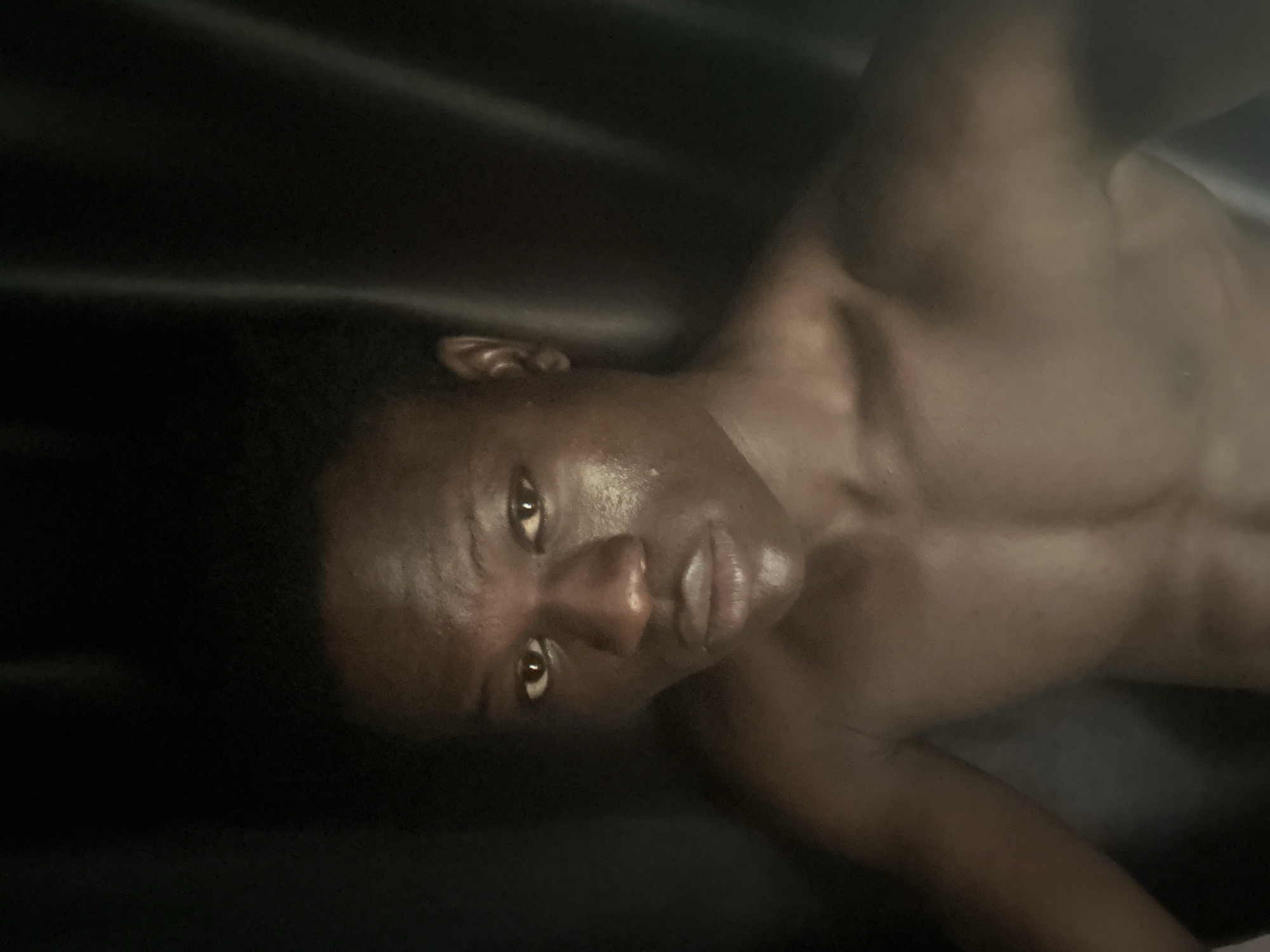 The image shows a close-up of a young Black male's face and upper chest, taken from a high angle. He has dark skin, short black hair, and appears to be looking directly at the camera. His face is illuminated by a light source, highlighting his facial features and casting shadows. The background is dark and appears to be a draped fabric, possibly a curtain or backdrop. There is no discernible urban environment visible in the image, and therefore, no specific location in Badagry, Nigeria can be determined. The image focuses solely on the individual.