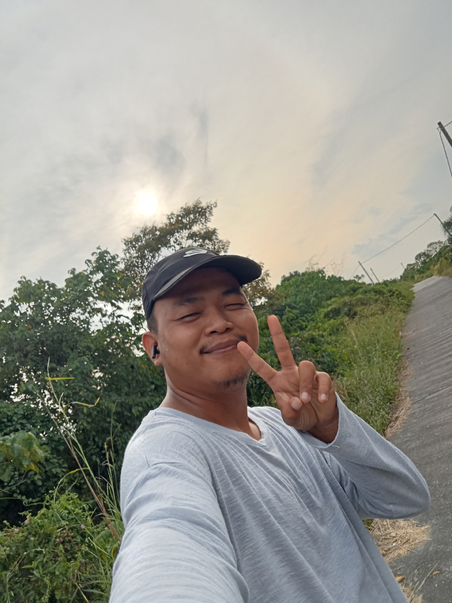 A man wearing a black baseball cap and a light gray long-sleeved t-shirt is posing for a selfie outdoors. He is smiling and making a peace sign with his right hand. He has a small mustache and goatee and is wearing a black wireless earbud in his left ear. Behind him, there is lush green vegetation and trees. A paved path or road slopes upwards to the right, with power lines visible in the distance. The sky is overcast with the sun shining through the clouds. The overall scene suggests a rural or semi-rural setting, possibly on the outskirts of Teluk Kenidai, Indonesia.