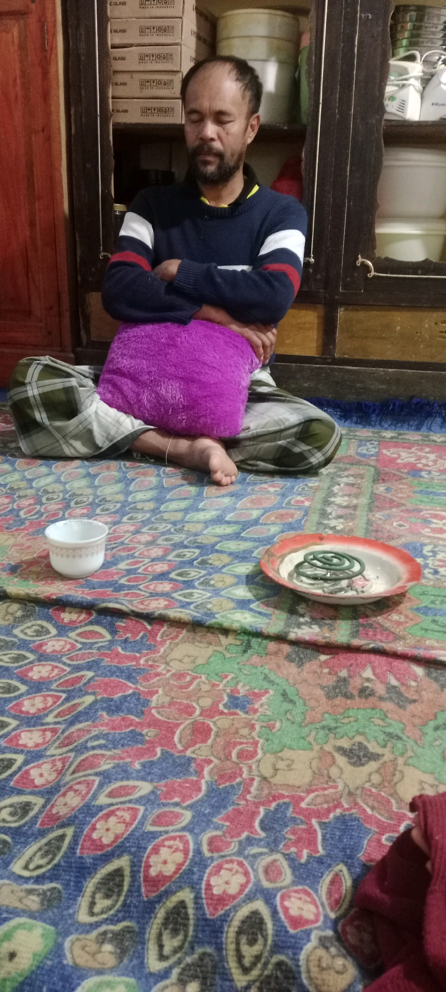 A man is sitting cross-legged on a patterned rug. He is wearing a dark blue sweater with white and red stripes on the sleeves. He has his arms crossed and his eyes are closed. A purple pillow is placed on his lap. To his left, there is a small white bowl. To his right, there is a shallow red and white dish containing a coiled mosquito repellent. The background shows a wooden cabinet with shelves containing various items. The scene appears to be indoors, possibly in a home in Kassi Buleng, Indonesia.