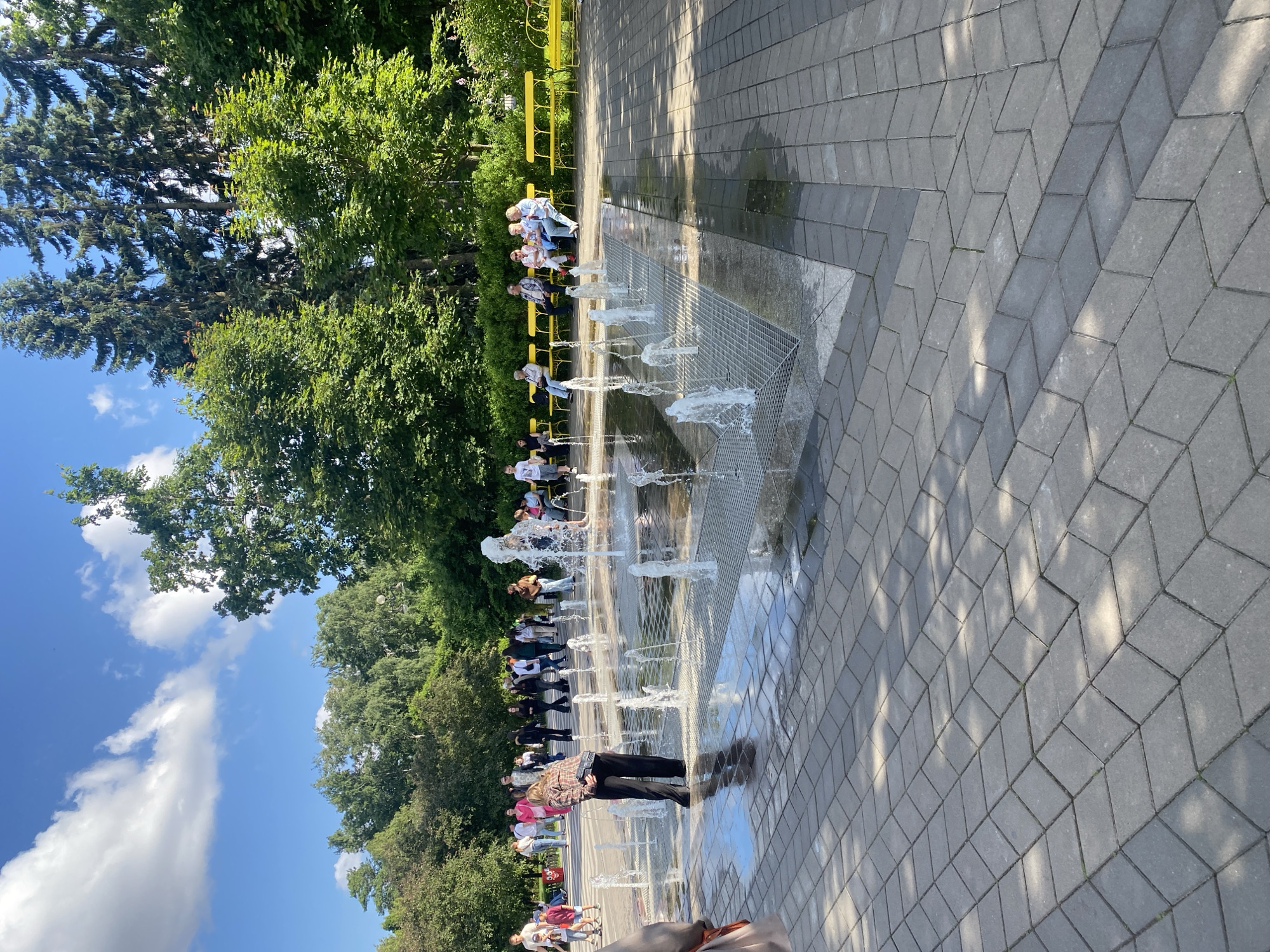 This image depicts a lively public square in Saint Petersburg, Russia, likely during warm weather. In the foreground, a modern splash pad with multiple water jets is active, with water spraying upwards from the pavement. Several individuals are seen interacting with the water, including a woman in the center foreground bending over, and a few other figures in the background. Surrounding the splash pad, a paved area with a distinctive geometric pattern of grey interlocking stones is visible. Beyond the paved area, bright yellow benches are arranged, occupied by groups of people relaxing and observing. The background is characterized by lush green trees and a clear blue sky with scattered white clouds, suggesting a park-like or recreational area within the city. The overall atmosphere appears to be one of leisure and enjoyment.