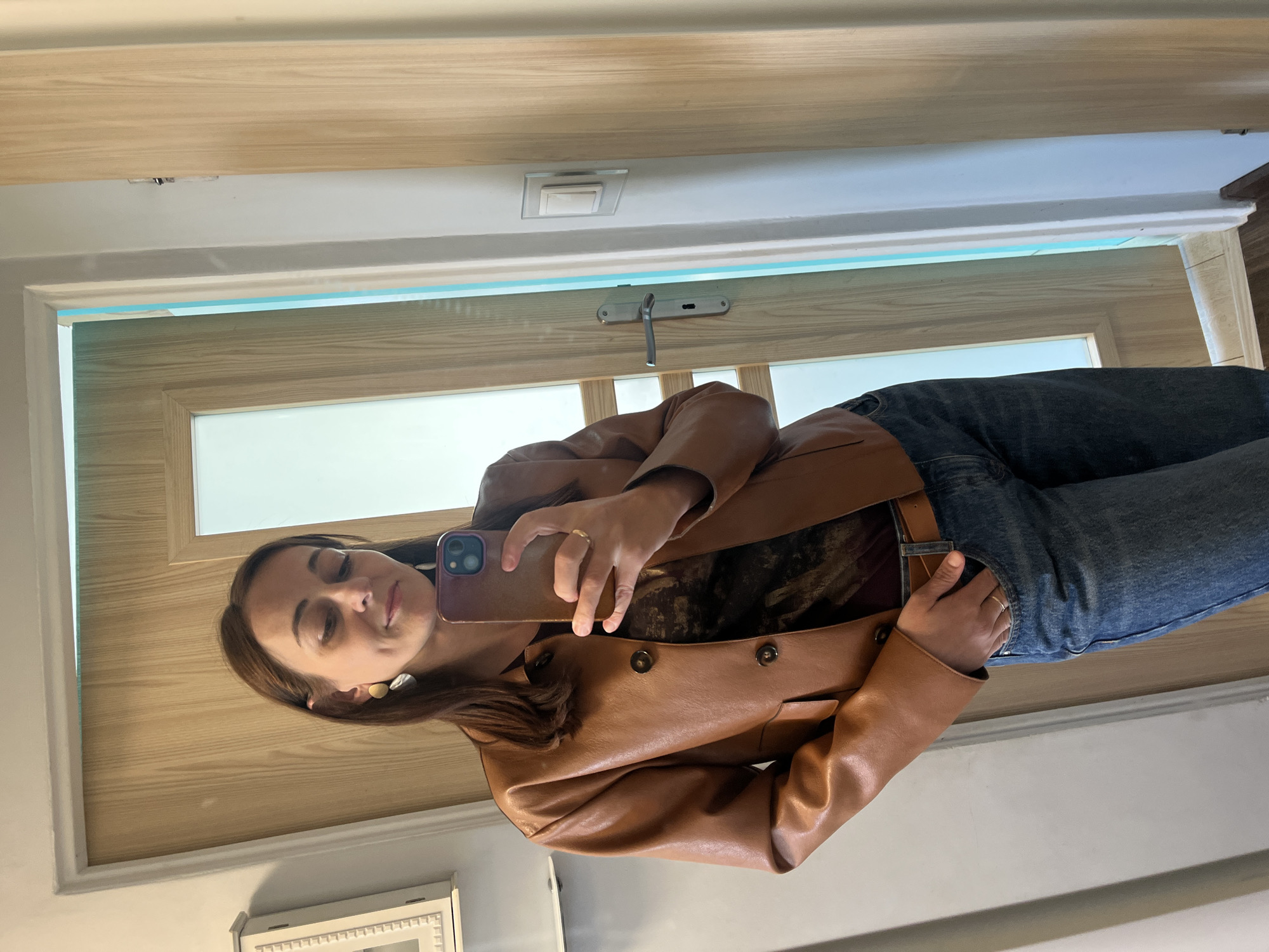 A woman stands indoors, taking a selfie in a mirror. She has brown hair and is wearing a brown leather jacket, a patterned shirt, blue jeans, and a brown belt. She is also wearing large, unique earrings and a ring. The background shows a wooden door with a frosted glass panel, and a light switch on the wall. The floor is made of wood.