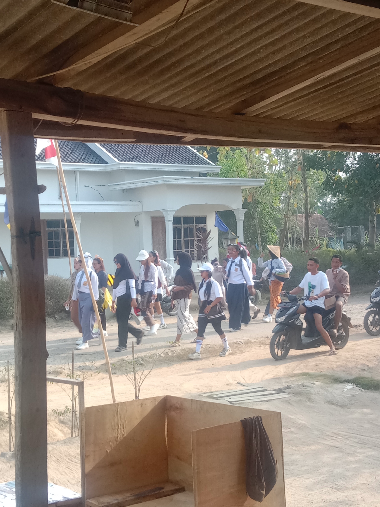In a dusty outdoor scene, a group of individuals, predominantly young women in school uniforms, walk along a dirt road in front of a white building. The building has a tiled roof and several windows. Some individuals are wearing headscarves, and at least one person is wearing a conical hat. A man and a passenger are riding a black motorcycle on the right side of the road. Another motorcycle is visible in the far right background. The foreground shows a wooden structure with a draped cloth. The environment appears to be rural or semi-urban, with trees and vegetation visible in the background. The sky is clear and the lighting suggests it is daytime.