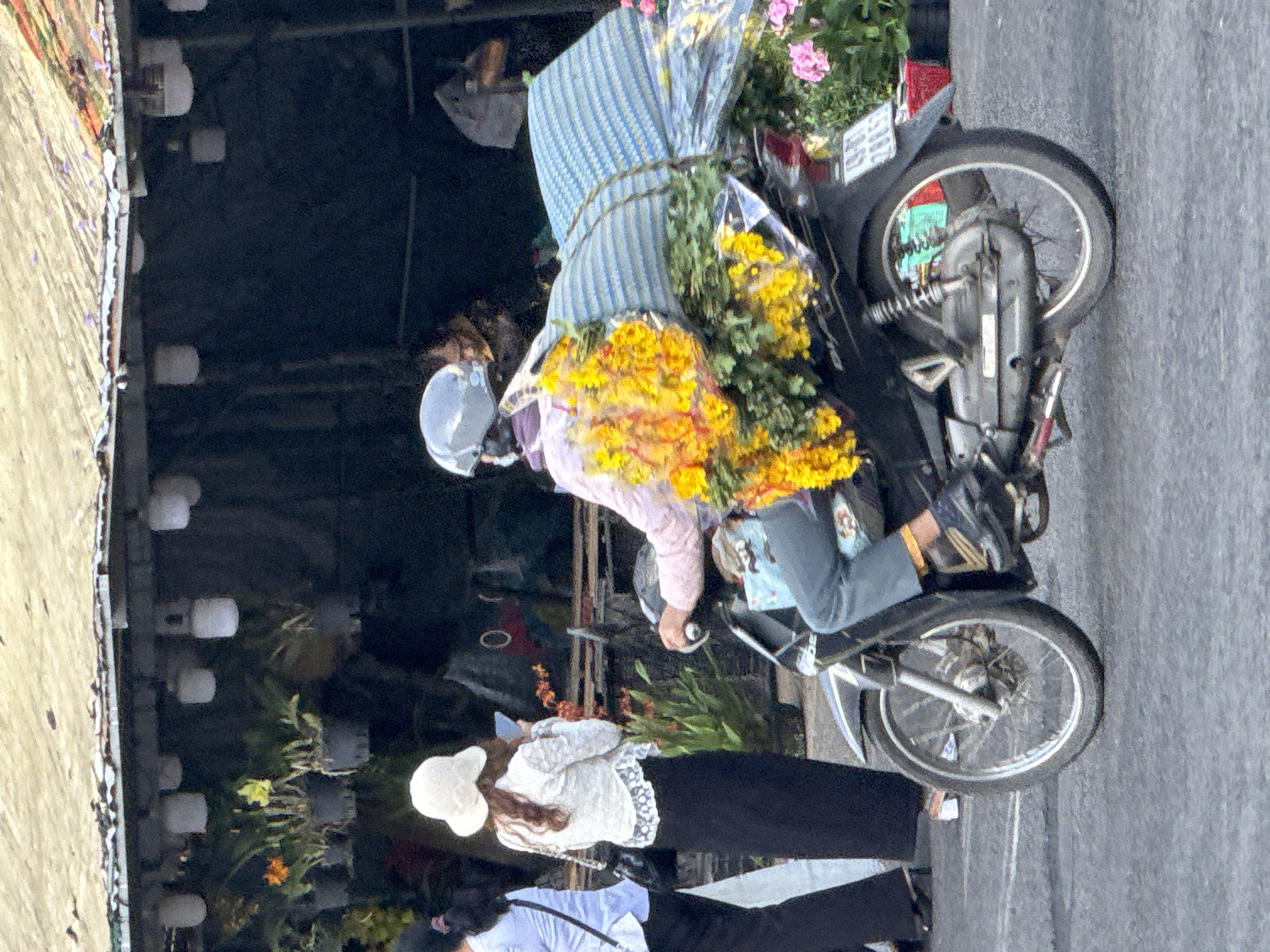 A person wearing a helmet and a pink shirt rides a motorcycle, carrying a large bundle of yellow flowers on the back. The motorcycle is dark colored with silver accents and has a license plate with the numbers 49-B1 288.54. The person is wearing blue pants and black shoes. In the background, there is a market stall with various plants and flowers. A woman in a white hat and white top with black skirt is standing in front of the stall, looking at her phone. Another person in a white shirt and black pants is to the left of the woman. The scene appears to be in Da Lat, Vietnam, likely a flower market, given the abundance of flowers and the presence of market stalls. The ground is asphalt.