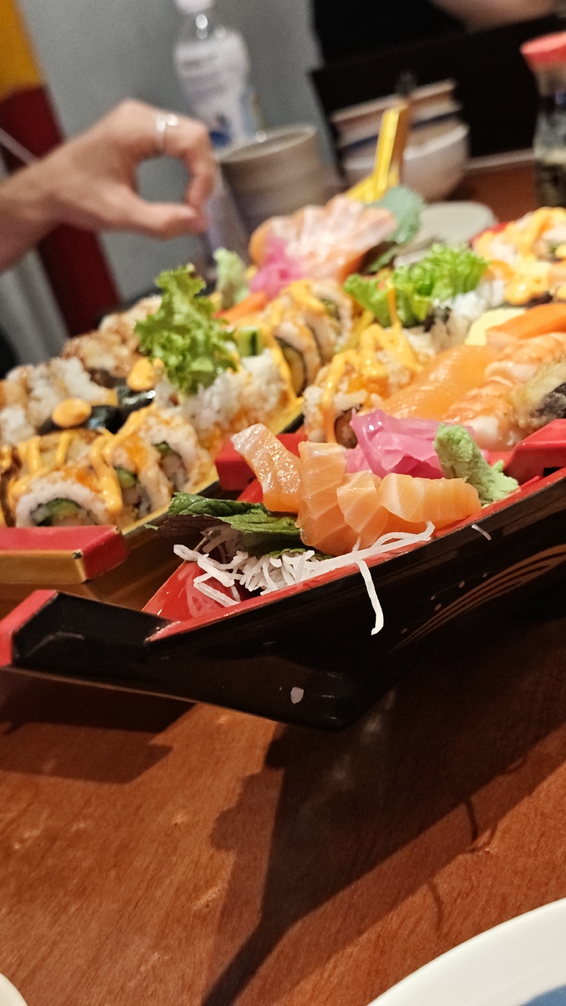 A close-up, low-angle shot depicts an elaborate platter of sushi and sashimi, resting on a dark wooden table. The sushi is arranged in a boat-shaped container, lined with red accents and intricate details. Various types of sushi rolls are visible, topped with sauces, lettuce, and shredded vegetables. Slices of salmon sashimi, pickled ginger, and wasabi are also present.
A hand wearing a ring is partially visible in the background. Behind the platter, there are blurred objects including a plastic water bottle, a cup, and bowls. The background suggests an indoor restaurant setting. The image likely originates from Đà Nẵng, Vietnam, based on the question's context.