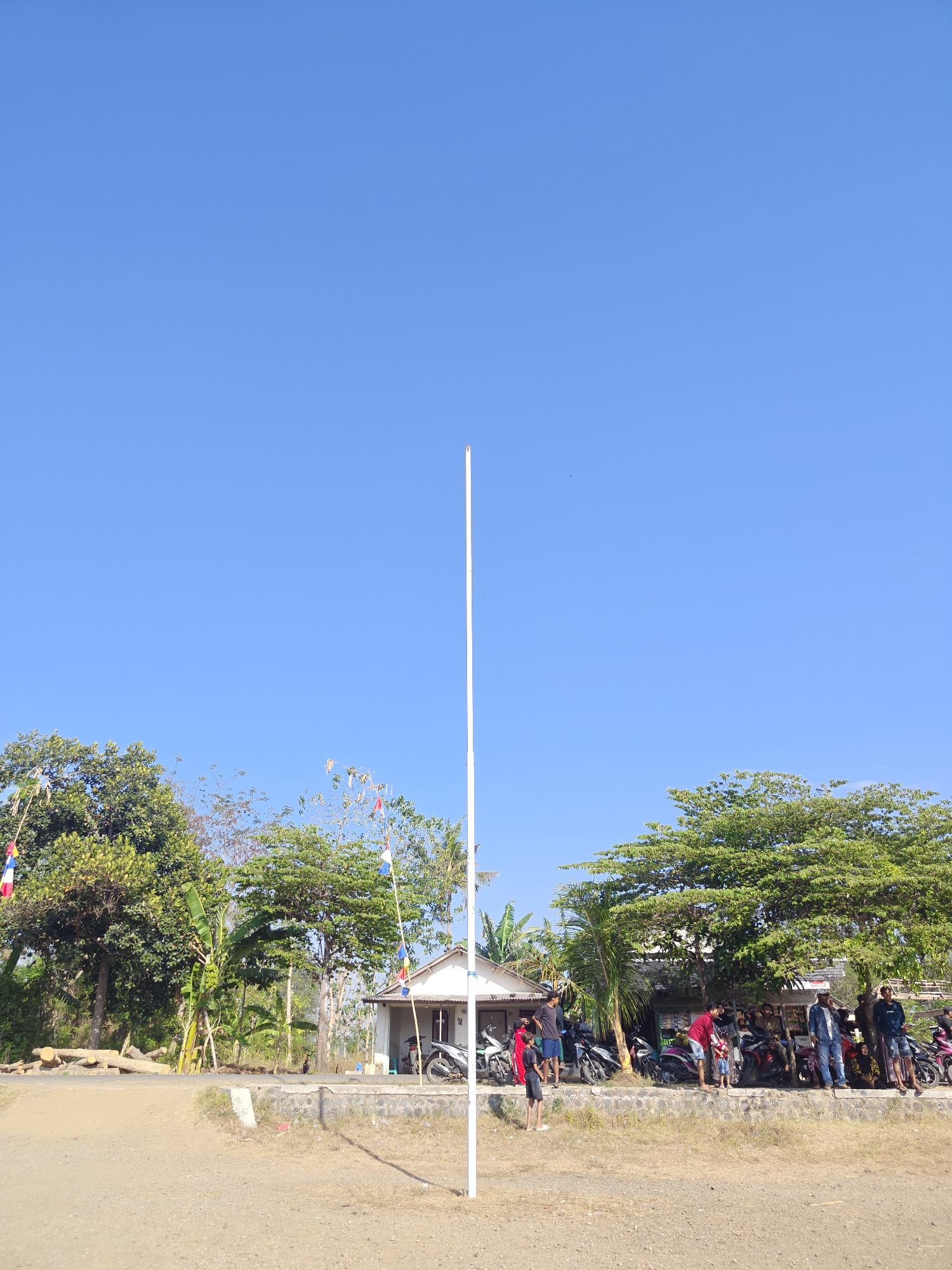The image depicts a scene in Cermee, Indonesia, under a clear blue sky. A tall, white pole stands prominently in the foreground, extending towards the sky. To the left of the pole, a group of trees with lush green foliage dominates the mid-ground, with a small, white building situated behind them. A few banana trees are also visible amongst the larger trees. In front of the building, several motorcycles are parked, and a small group of people are gathered. Further to the right, more trees provide shade, and another collection of people and motorcycles are present. The ground in the foreground is a dry, dusty dirt path, and a low stone wall runs horizontally across the mid-ground. Small flags with colorful designs are strung between poles in various locations around the scene. The overall impression is of a rural or semi-rural setting, possibly a gathering or event taking place outdoors.