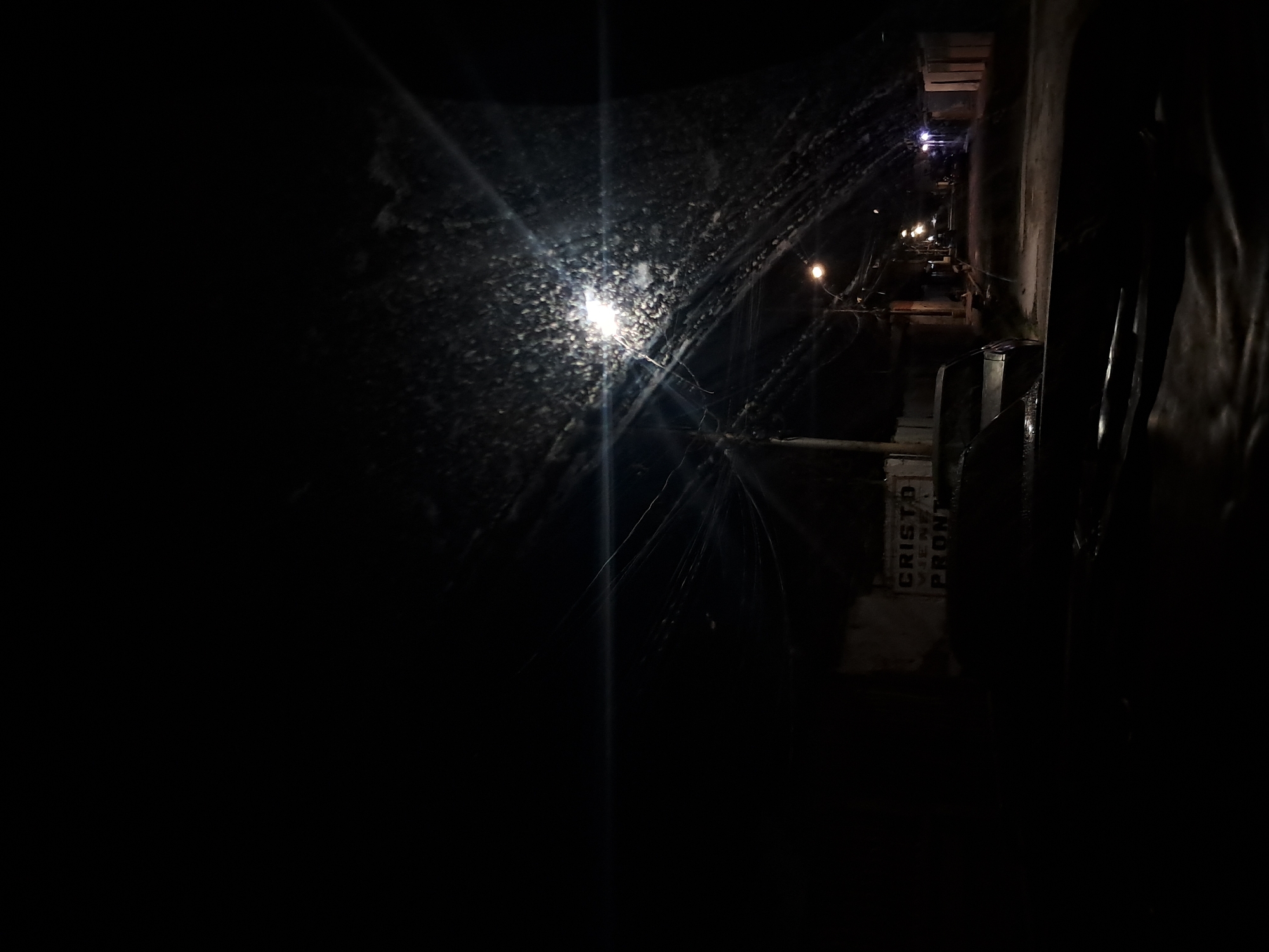 A street scene in Cumana, Venezuela at night. The image is dark, with a bright street lamp in the upper right quadrant illuminating a gritty, textured surface, possibly a window or windshield covered in water droplets or grime. Several power lines crisscross the scene, and another, dimmer street lamp is visible further down the road. The road itself is paved and appears wet. In the foreground, the hood of a vehicle is visible. To the left, a wall with writing on it is partially obscured by darkness, with the words "CRISTO VIENE PRONTO" (Christ is coming soon) discernible. The scene depicts a typical urban street at night, with hints of urban decay and religious messaging.