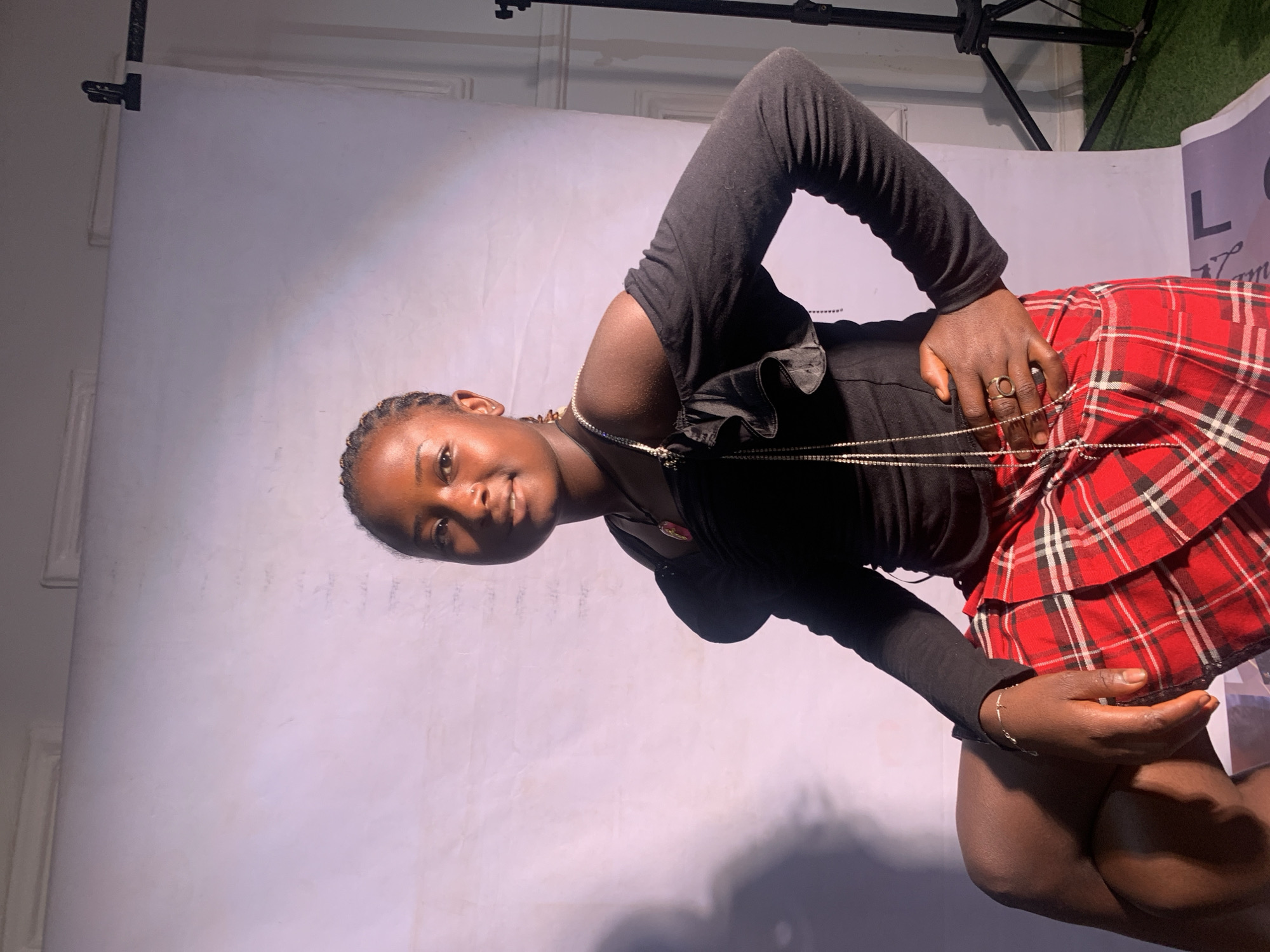 A person is photographed indoors against a backdrop. The individual is wearing a black long-sleeved top and a red plaid skirt. A gold necklace adorns their neck, and rings are visible on their fingers. The location and city of Jos, Nigeria, are not confirmed through the image's visual elements. The photo may be part of a studio photoshoot.