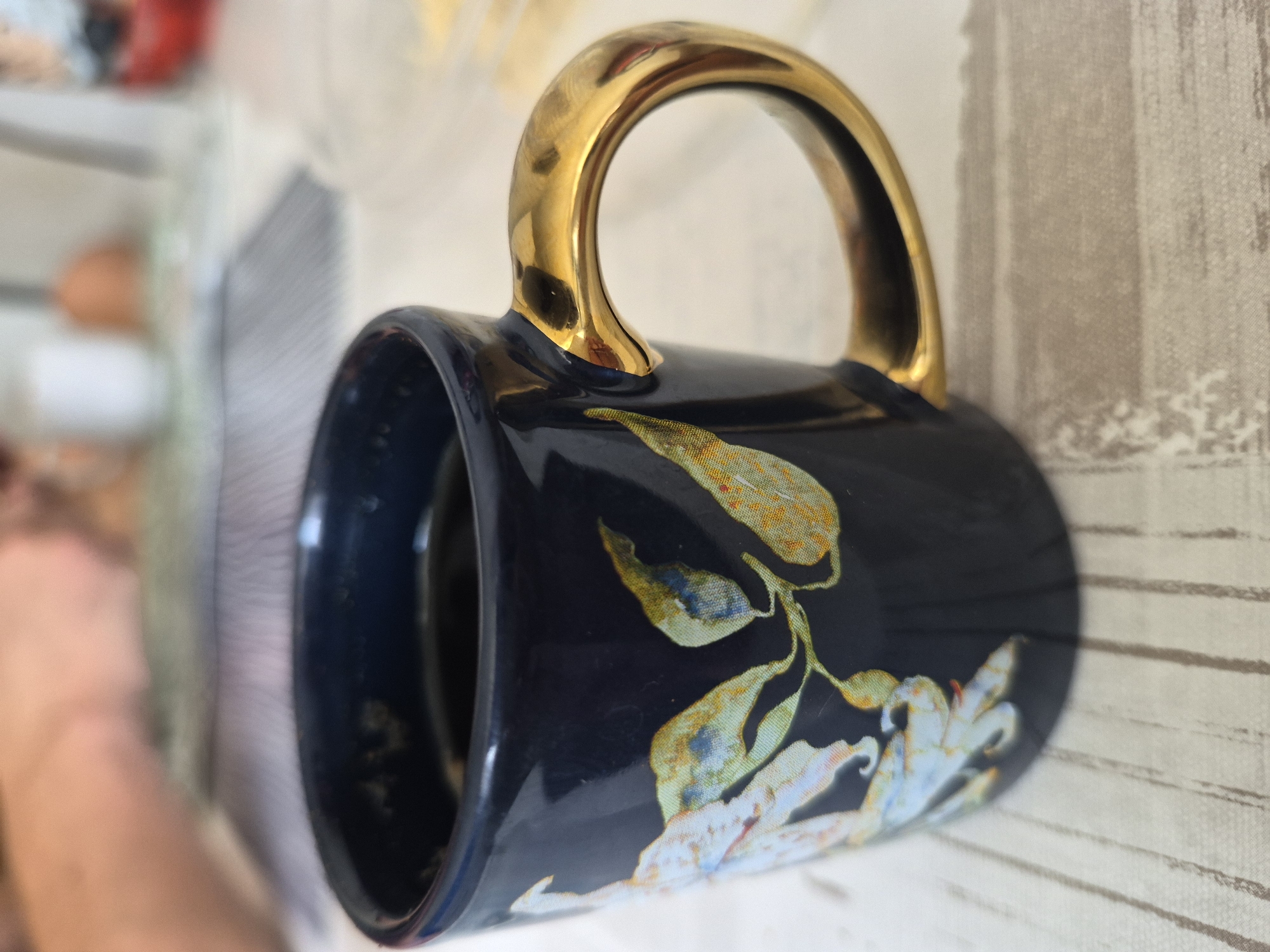 A close-up, eye-level shot focuses on a dark blue ceramic mug with a gold handle, resting on a light-colored, horizontally striped surface. The mug features a floral design in muted tones of white, yellow, and blue. The surface it sits on appears to be a table or placemat. In the blurred background, hints of human activity are visible, including what looks like hands and other objects, suggesting a domestic or casual setting.