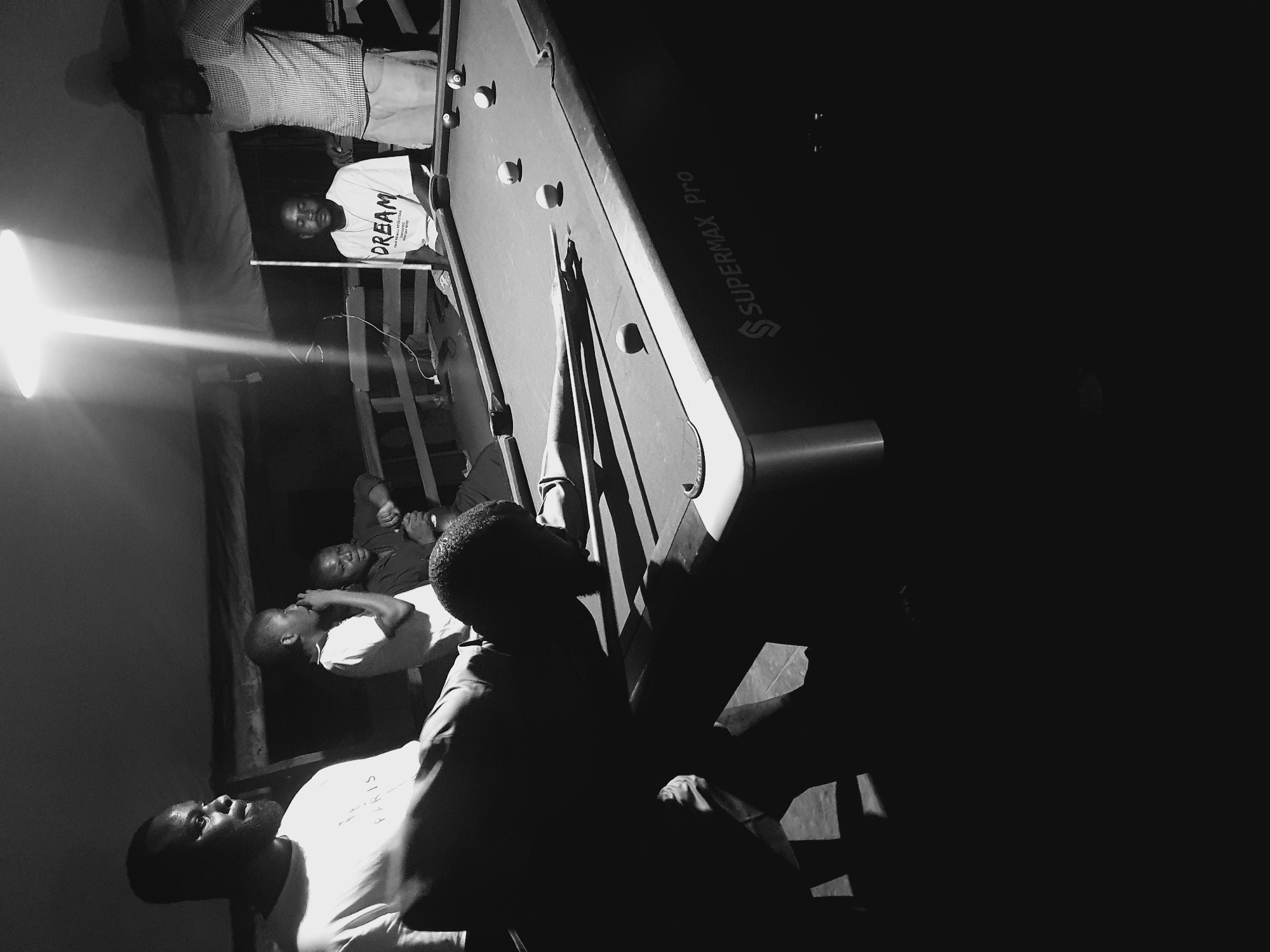 In Lokoja, Nigeria, a group of men are gathered around a pool table in a dimly lit indoor setting, possibly a local bar or recreational area. A bright overhead light illuminates the pool table and casts strong shadows. One man, positioned in the foreground, is bent over the table in a pool-playing stance, his face obscured by shadow. Other men are visible in the background, some standing and some seated. One man wears a t-shirt with the word "DREAM" printed on it. The pool table is set up with several balls. The overall atmosphere appears casual and social.