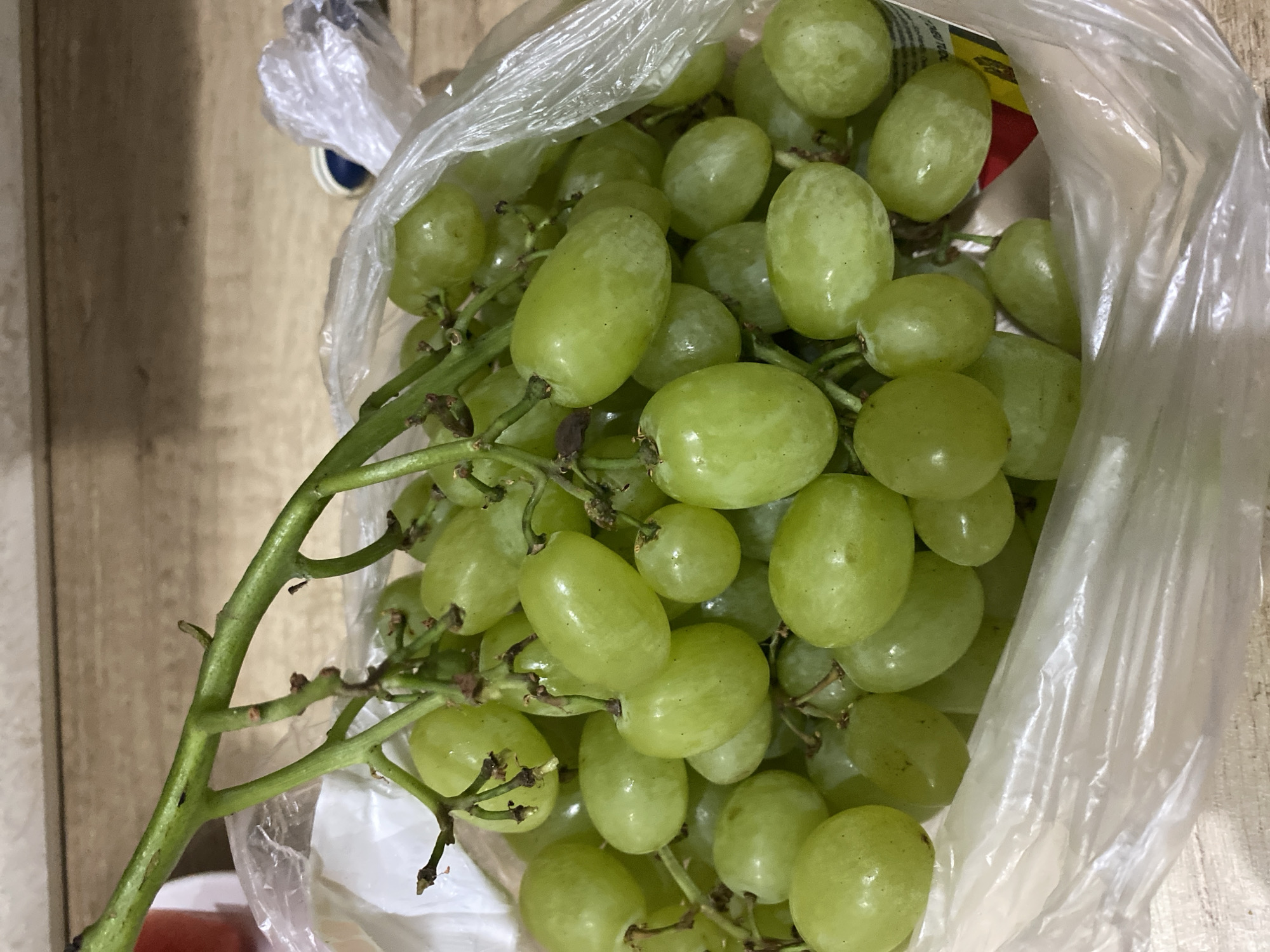 A bunch of green grapes is presented in a clear plastic bag, resting on a wooden surface. The grapes are plump and have a smooth, translucent skin, with some subtle variations in color from pale green to a more vibrant hue. They are attached to thin, green stems, forming a clustered arrangement. The plastic bag is slightly crinkled, partially obscuring some of the grapes and creating a reflective surface. The wooden surface beneath appears to be a table or countertop with visible grain patterns. The background is softly blurred, suggesting an indoor setting. There are no people present in the image. The context provided about Chisinau, Moldova, is not visually discernible from the image itself.