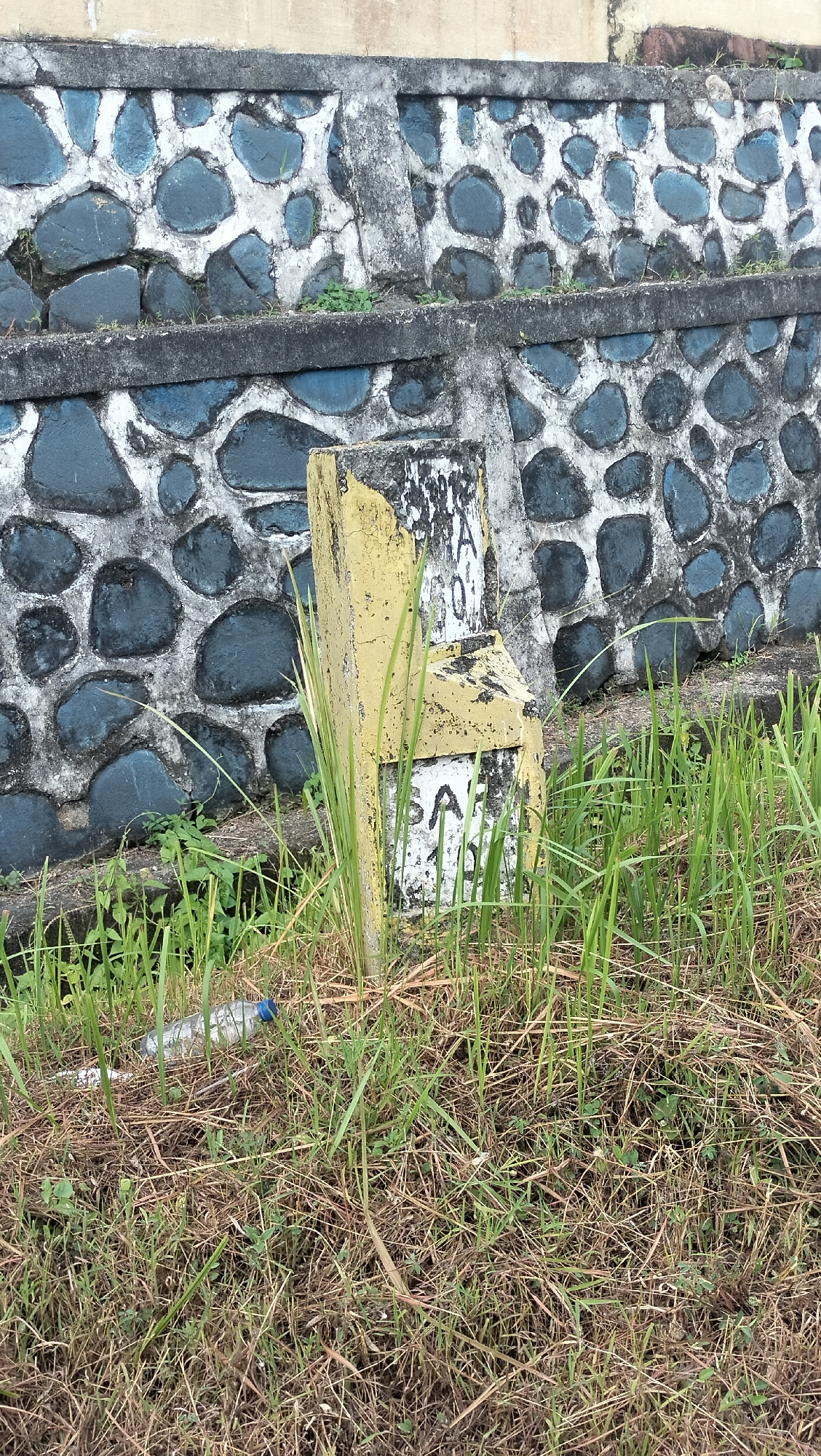 This full-shot image captures an outdoor scene in Lamtamot, Indonesia, featuring a weathered stone wall and a partially broken marker post in the foreground. The wall, likely a retaining wall or barrier, is constructed from dark, irregularly shaped stones embedded in light-colored concrete or mortar, with a prominent horizontal concrete beam dividing its sections.
In front of the wall, a fractured concrete or stone marker stands amidst grass. The marker is predominantly yellow, though much of its paint is chipped and faded, revealing a white or light gray base. Text is partially visible on its surface; the upper fragment shows characters such as "...A 0" or "...IA 0", while the lower part clearly displays "SA" followed by what appears to be a "0" or "O" below it, though the full inscription is unreadable.
The ground around the marker and in the foreground is covered with a mix of green grass and dry, brown vegetation. A discarded clear plastic water bottle with a blue cap lies on the ground amidst the grass to the left of the marker. The scene is illuminated by natural daylight, suggesting it is daytime. There is no visible activity or people, presenting a static view of an aged and somewhat neglected area, possibly roadside or adjacent to a structure.