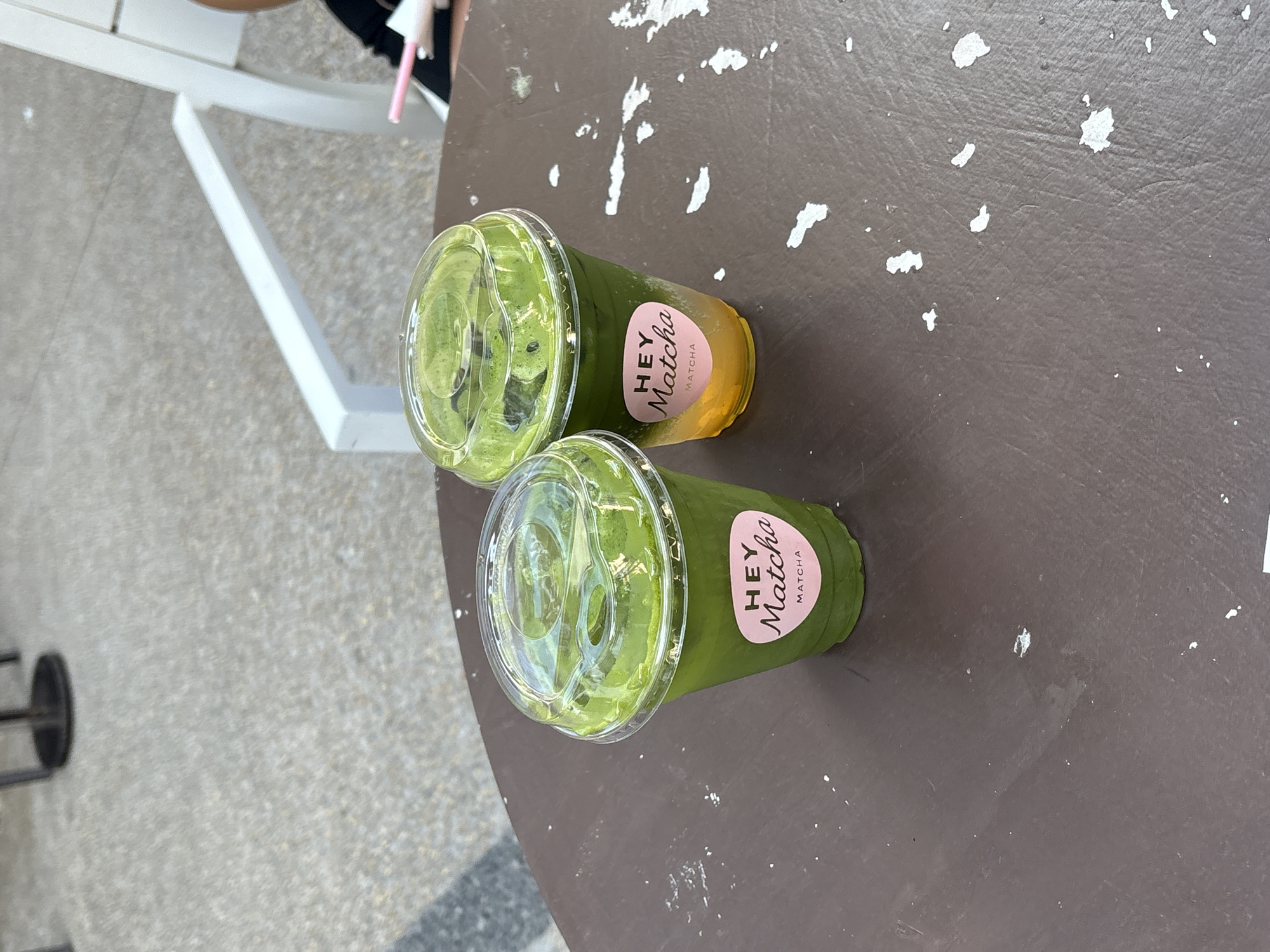 Two cups of green matcha drinks with clear plastic lids and "Hey Matcha" stickers are on a dark brown table. The table has white specks and wear. Part of a white chair is visible in the upper left of the image. The location is San Bernardino Tlaxcalancingo, Mexico.