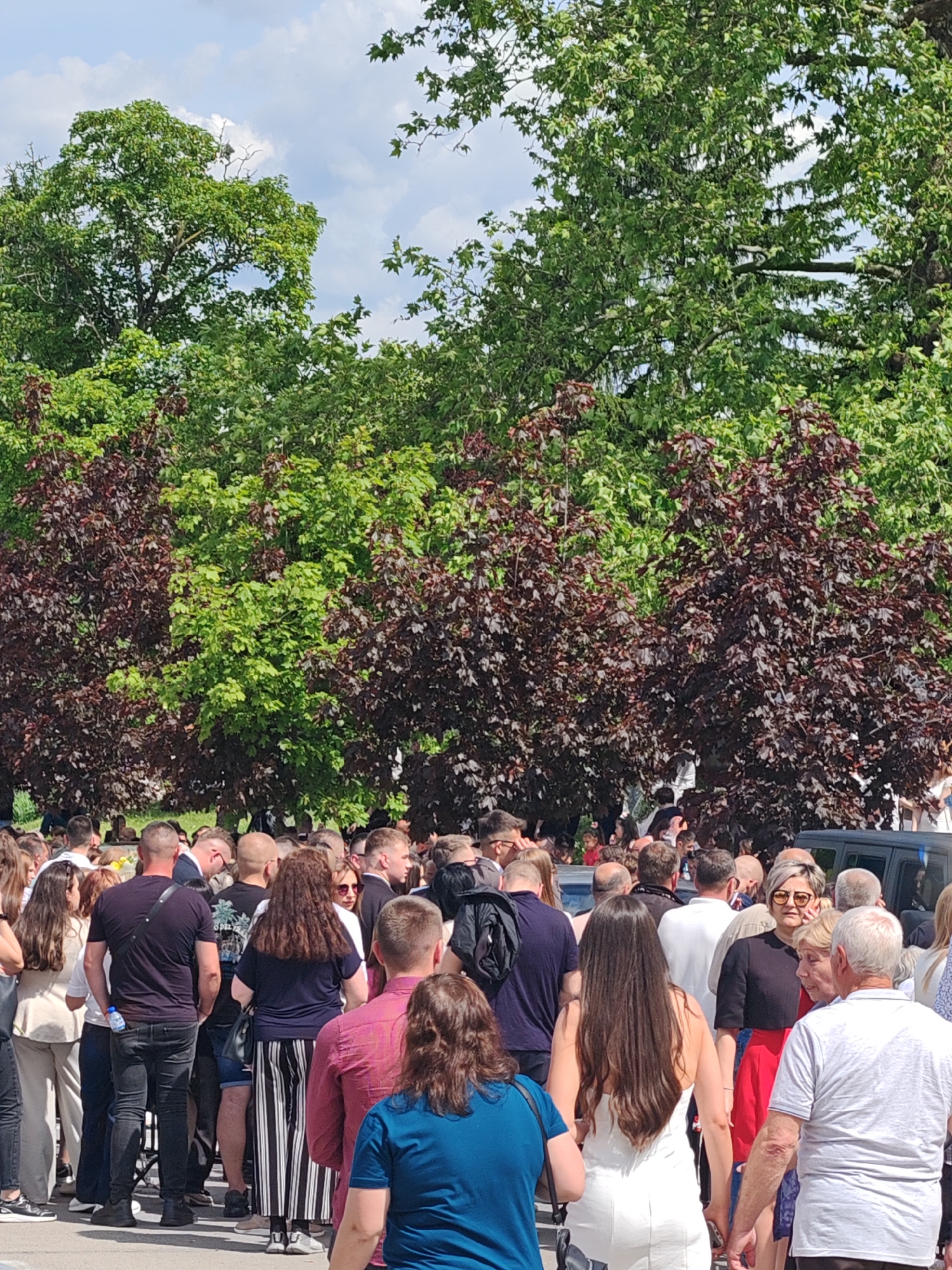 The image shows a crowd of people gathered in an outdoor location. The crowd seems dense, with people standing close to each other. A mix of men and women are visible, dressed in various casual clothing. The background consists of green and maroon-leaved trees. A vehicle is partially visible in the right background. Based on the presence of crowds, the image is likely taken in the city of Shumen, Bulgaria during a public event or gathering.