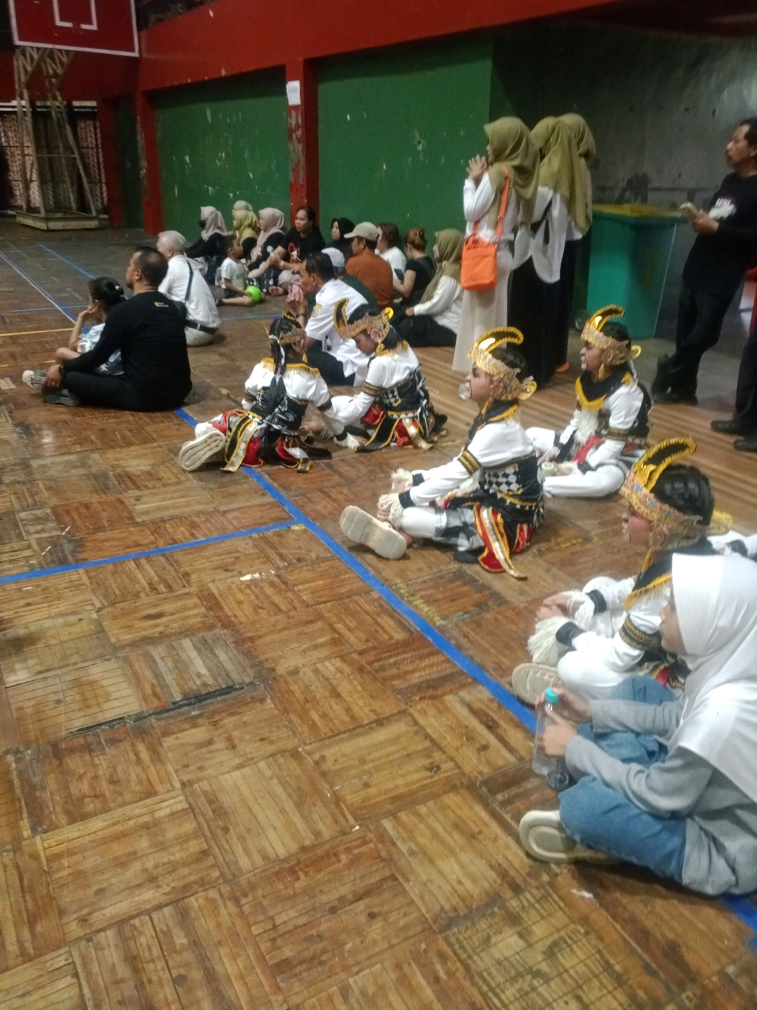 In a gymnasium with a wooden floor marked by blue lines, a group of people are gathered, seated on the floor. In the foreground, several children are dressed in traditional attire, complete with elaborate headdresses. One child in the far right foreground, wearing a white hijab and grey top, is holding a plastic water bottle. Behind them, adults are seated, some watching the children. In the background, there is a basketball hoop and a green wall. To the right, a man in a black shirt is standing. The scene appears to be in Wuryorejo, Indonesia, likely during a cultural performance or event.