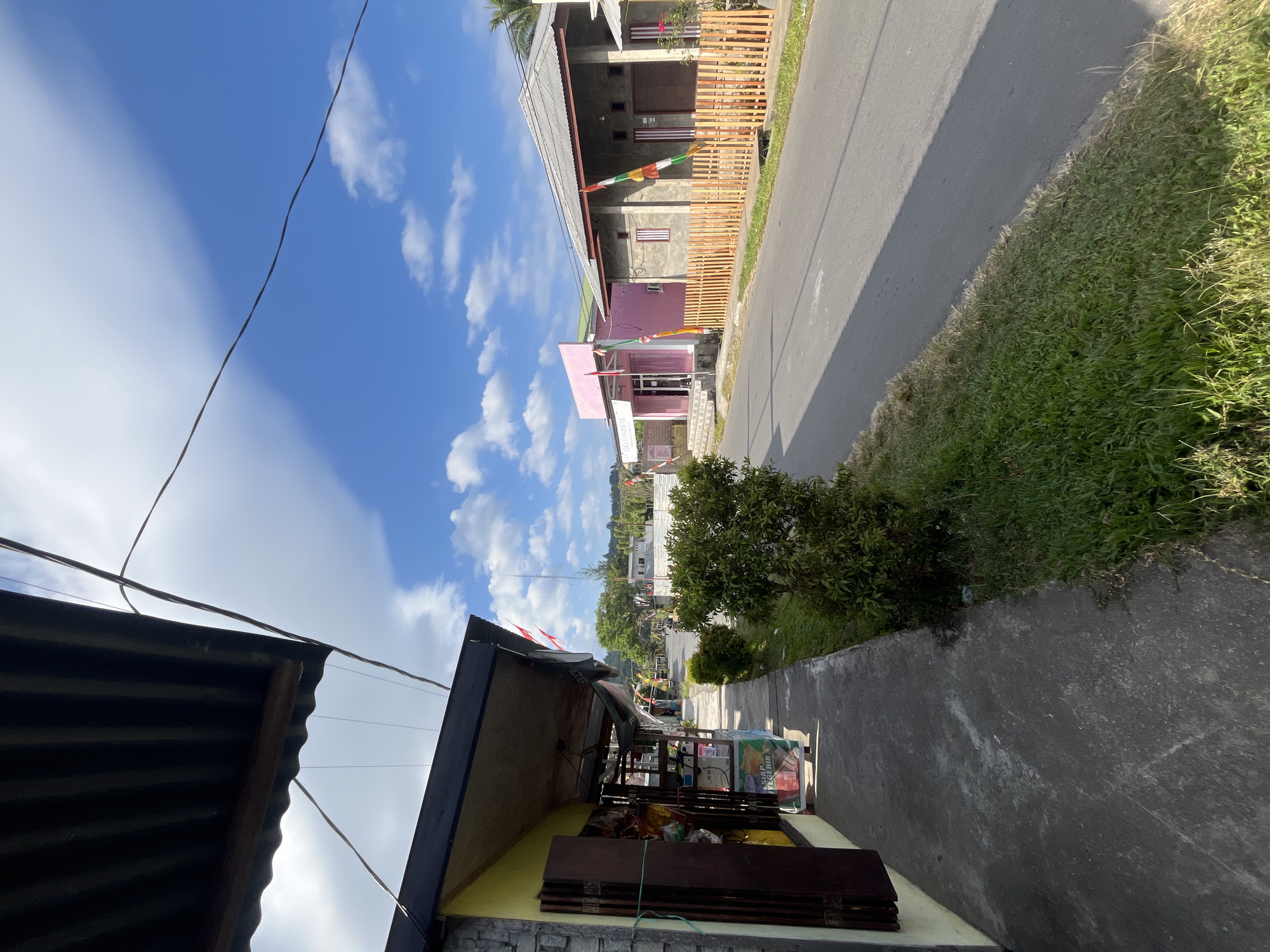 The image captures a street scene in Sanana, Indonesia, on a bright, sunny day. The foreground shows a sidewalk and a narrow grassy strip next to a paved road. To the left, a small shop with a corrugated metal awning and yellow-painted walls is visible, displaying various goods on shelves outside. The shop has dark wooden shutters that are partially open, revealing products. Further down the street, a pink building with signage that reads "Choco Coshop" is present. Adjacent to it is a gray house with a gray, sloped roof, featuring a light orange wooden fence in front. Several Indonesian flags, red and white, are displayed on poles along the street and on the fence. The sky is blue with scattered white clouds. The environment suggests a residential or mixed-use area in a town or city. No people are clearly depicted in the scene.