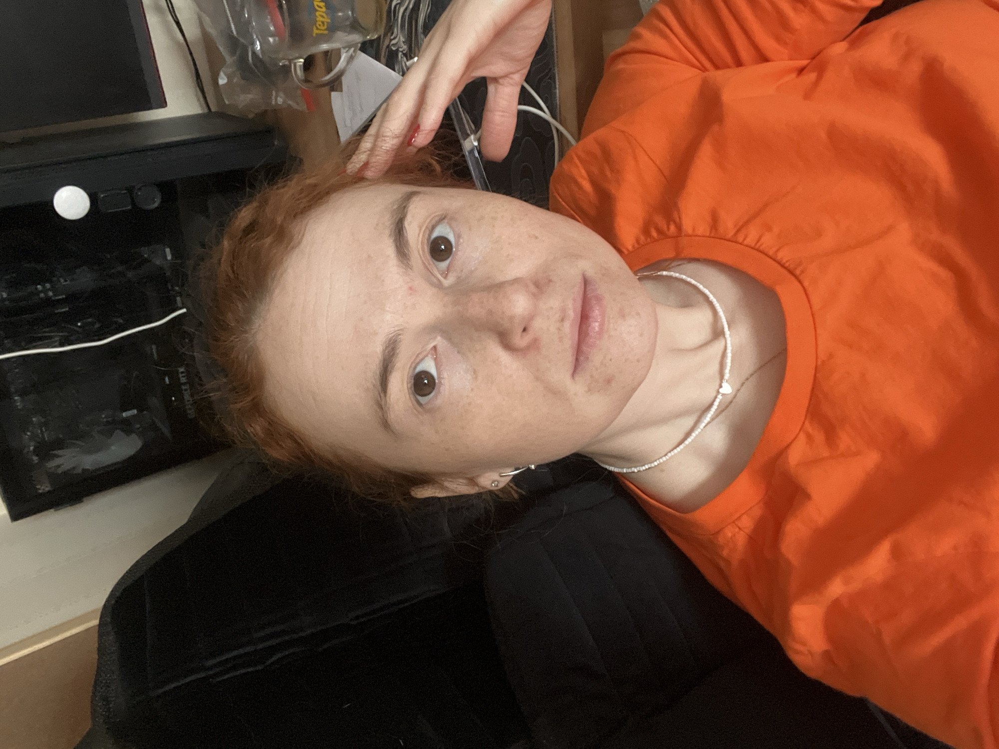 The image presents a close-up, rotated portrait of a woman in an indoor setting, likely in Chaikovsky, Russia. She has reddish-brown hair, fair skin with noticeable freckles, and dark, wide-open eyes fixed on the camera. She is wearing an orange long-sleeved top and a white beaded necklace with a small heart charm. A silver hoop earring is visible in her left ear. Her left hand, adorned with red nail polish, is raised and positioned near her head. Her expression appears neutral or slightly curious.
The background shows electronic equipment, including a black computer tower with a visible white circular power button and other controls. Faint text "GEFORCE RTX" is discernible on the tower. A monitor screen is partially visible above it, along with various white cables. A clear glass or mug, hanging upside down, features a yellow label with the partial text "Tepa". The overall scene suggests a casual, personal environment. No specific activity is taking place beyond posing for the photograph. The time of day is not discernible from the indoor lighting.