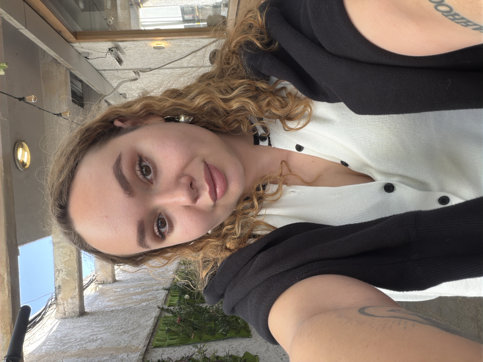 A young woman with curly hair is taking a selfie. She is wearing a white cardigan with black buttons and a black sweatshirt. Her skin tone is fair, and she has light brown eyes. A silver earring is visible in her left ear. A tattoo can be seen on her upper arm. The background appears to be an outdoor patio area with string lights and greenery. Given the context of Tel-Aviv, Israel, the architecture visible in the background suggests the image was taken in a neighborhood with a Mediterranean architectural style.