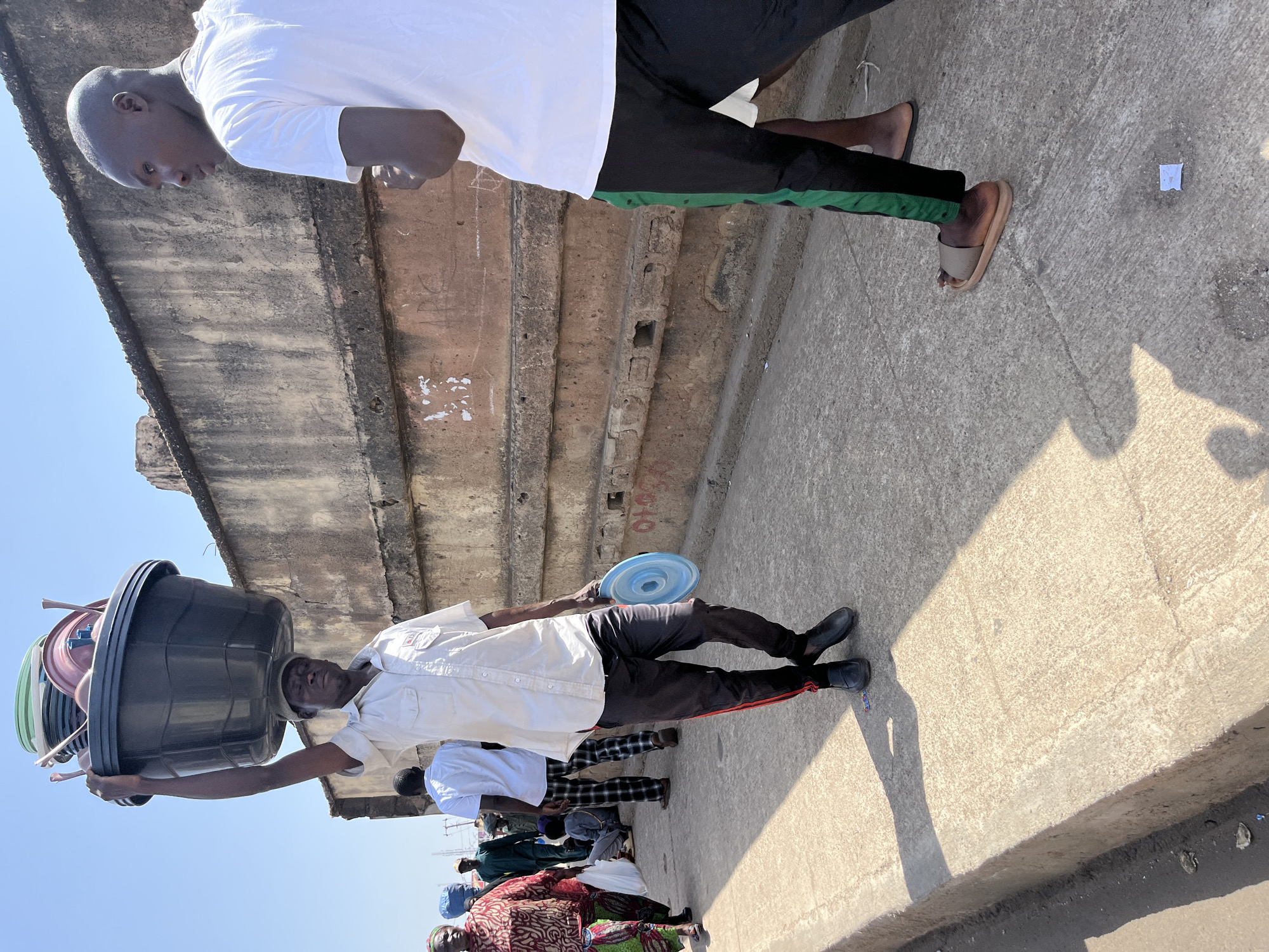 A street scene in Jos, Nigeria. An older man balances a large black tub full of colorful plastic objects on his head. He is wearing a white button-down shirt, dark pants with a red stripe, and black shoes. To the right of the older man, a group of people are walking. One man wears a white shirt and dark pants with a green stripe. He is wearing sandals. A concrete structure is visible to the left. The ground is concrete. A small white piece of paper is on the ground. The sky is clear and blue. Shadows are cast on the ground.