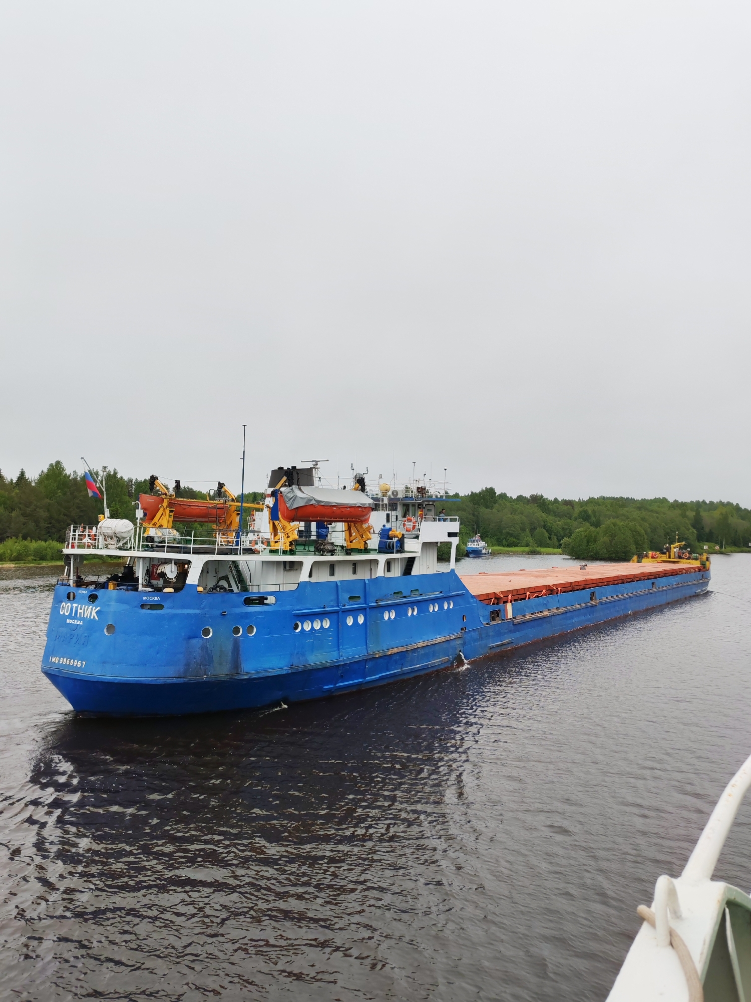 The image shows a blue barge named "Sotnik" sailing on a waterway, likely in the vicinity of Devyatiny, Russia, based on the barge's Moscow registration. The barge has an orange cargo hold and carries lifeboats, indicating it's equipped for passenger or crew safety. The sky is overcast, suggesting a cloudy day. Green trees line the banks of the waterway in the background. The perspective suggests the photo was taken from another boat.