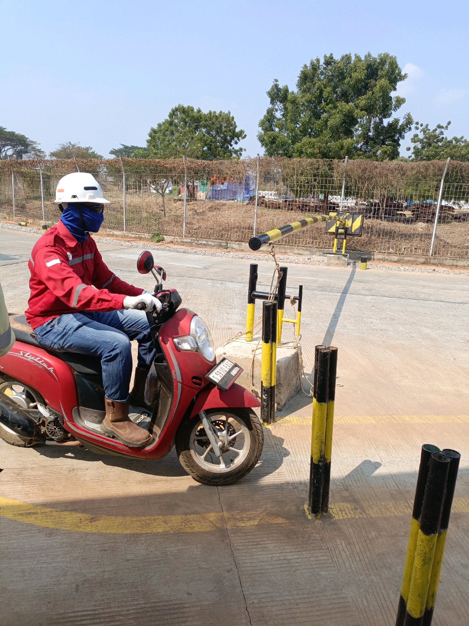 A person wearing a white hard hat, a blue face covering, a red long-sleeved work jacket with reflective strips, blue jeans, and brown boots is seated on a red scooter in Jenu, Indonesia. The person is holding the scooter's handlebars with white-gloved hands. The scooter has a license plate that reads "S 5910 FBO". In the background, a chain-link fence runs along the edge of a road, with trees and some construction materials visible beyond the fence. A yellow and black striped barrier arm is positioned across the road. Several yellow and black striped bollards are also present on the road. The ground is paved with concrete, and there are yellow painted lines on the surface. The sky is clear and blue, indicating a sunny day.
