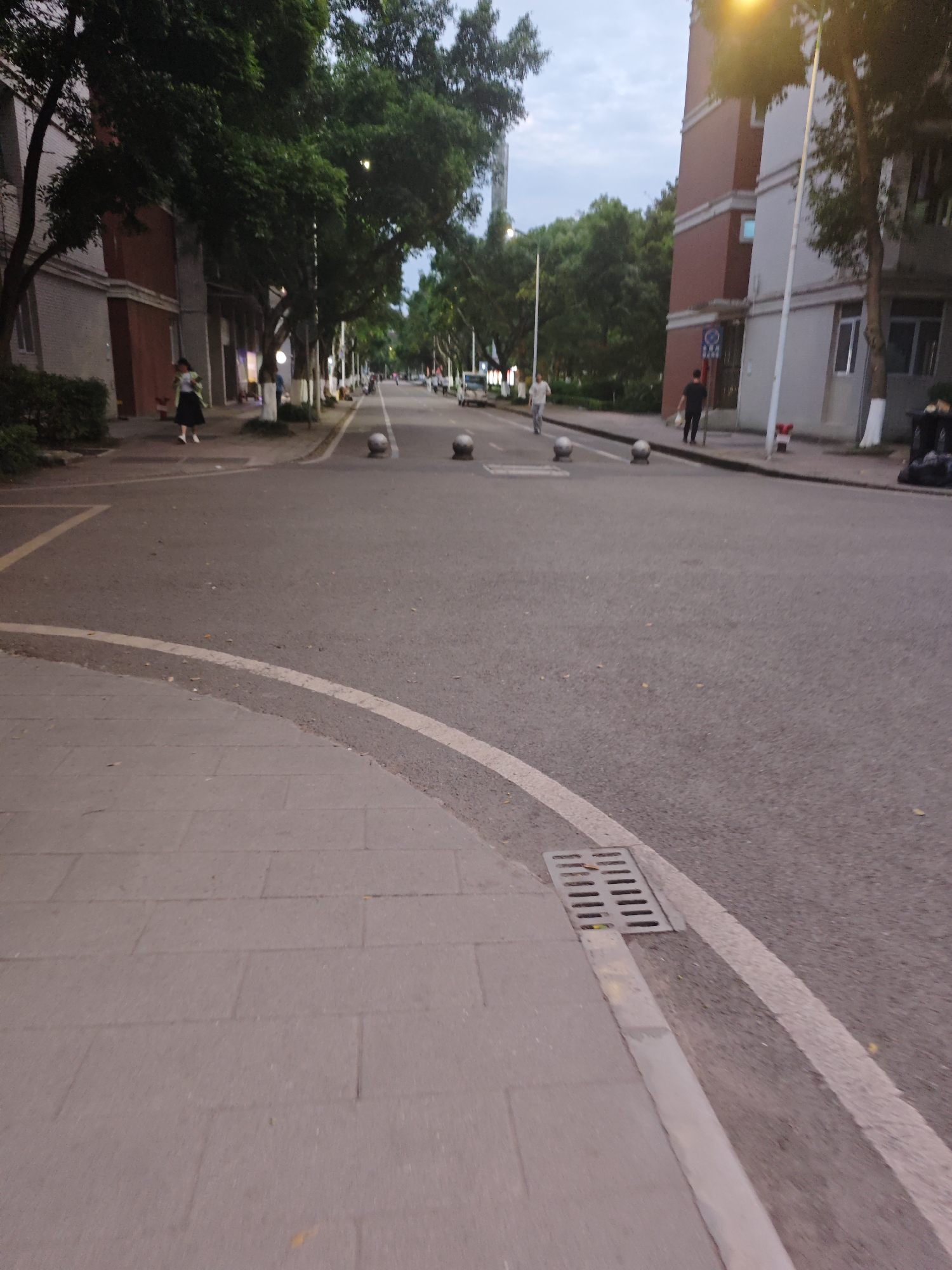 The image shows a street scene in Chongqing High-tech Zone, China. The street is paved with asphalt and lined with sidewalks made of gray rectangular paving stones. A white line separates the sidewalk from the road. A metal grate is visible on the edge of the sidewalk. Several round concrete bollards are positioned in the middle of the road. On either side of the road, there are buildings with red brick elements and green trees. Several people are walking on the sidewalks or the street. The lighting suggests it is dusk or dawn.