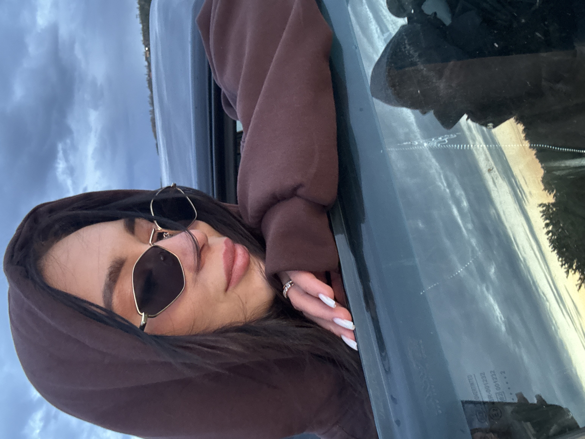 The image depicts a person leaning out of a dark-colored car window, resting on their side. The subject is a woman with long dark hair, wearing dark, angular sunglasses with thin gold-toned frames, and a dark brown hooded sweatshirt with the hood pulled up. Her head is tilted, with her cheek resting against the car's window frame or door. Her eyes appear closed or are looking downwards, and her lips are full and slightly parted. Her left hand is visible, resting on the window frame, adorned with white, manicured nails and a ring on her ring finger.
The car's window, which occupies the right side of the frame, reflects a hazy sky with hints of warmer light, possibly from a sunset or sunrise, above a dark, distant landscape featuring trees. The sky directly visible above the person's head is overcast with soft, grey clouds.
On the car window, several pieces of text are visible, appearing partially mirrored or inverted. These include the word "OVERINTED" and numerical codes such as "R-001232", "001232", and "E2". A dotted line, possibly indicating a heating element or antenna, is also present on the window surface. The overall impression is one of relaxation or contemplation, possibly during a scenic drive or stop, under either an overcast sky or during the early morning/late evening hours.